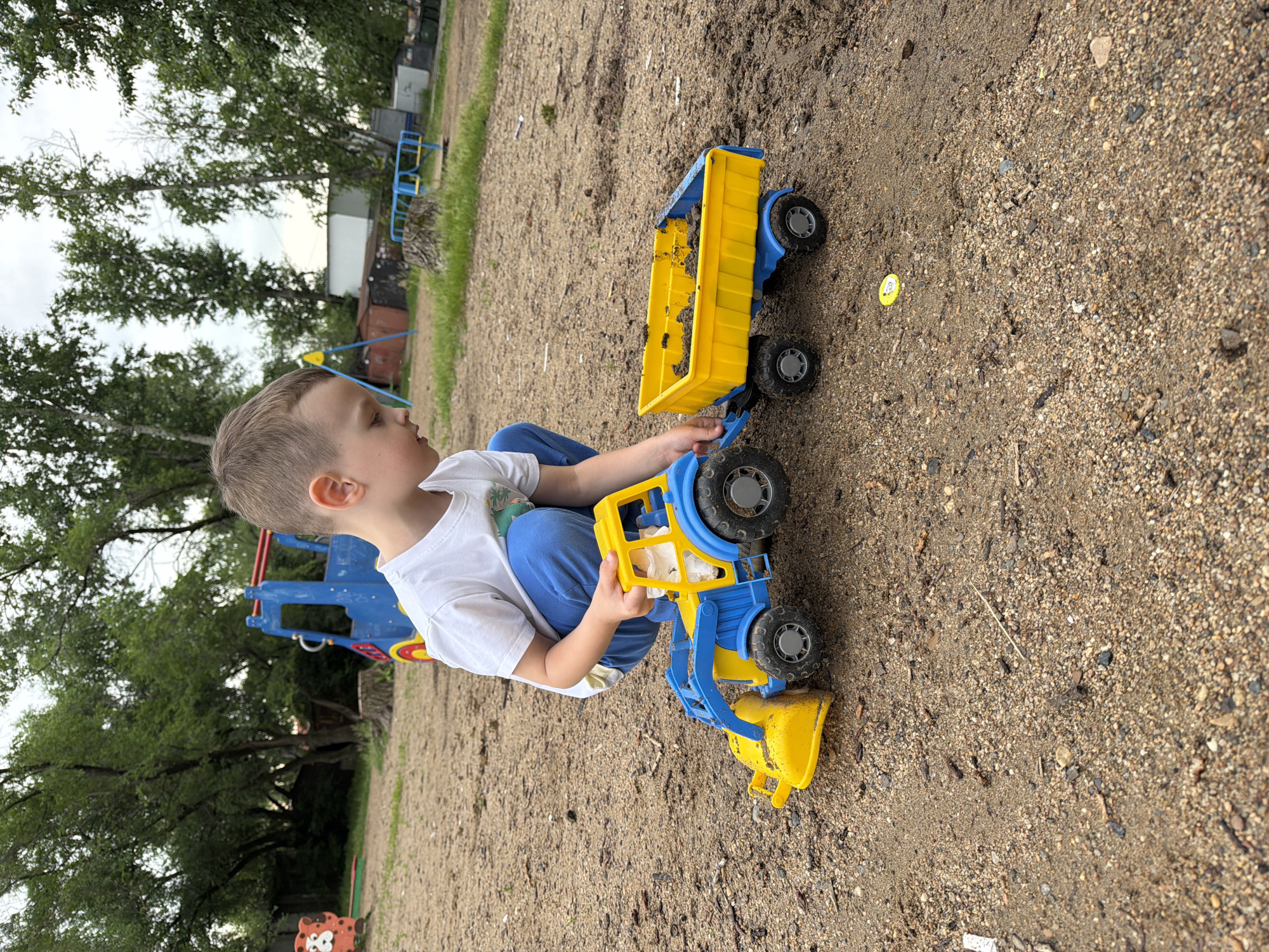 In a gravelly playground area, a young Caucasian boy, wearing a white t-shirt and blue pants, is crouched down, engaged in playing with a yellow and blue toy tractor and trailer. The tractor has a front-end loader filled with dirt, and the trailer is also loaded with dirt. The boy's right hand is on the tractor. In the background, there is playground equipment, including a blue structure with red accents and a red and orange object resembling a character. Trees with green foliage are visible behind the playground equipment. The ground is covered with small stones, dirt, and a few scattered twigs. The sky is overcast. The location is identified as a playground in Khabarovsk, Russia.