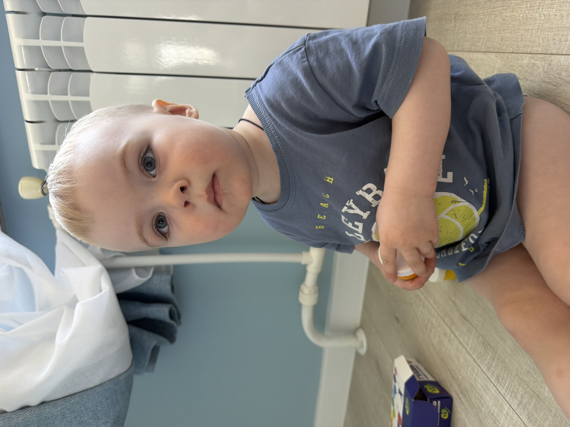A young child is sitting on a wooden floor in a room with light blue walls. The child is wearing a blue t-shirt with a lemon graphic and is holding a small container with yellow contents. To the left of the child are a white radiator and a pile of white and blue fabric. In the bottom right corner is a small box with blue and white packaging. Based on the environment, there is no evidence to confirm that the location is Votkinsk, Russia.
