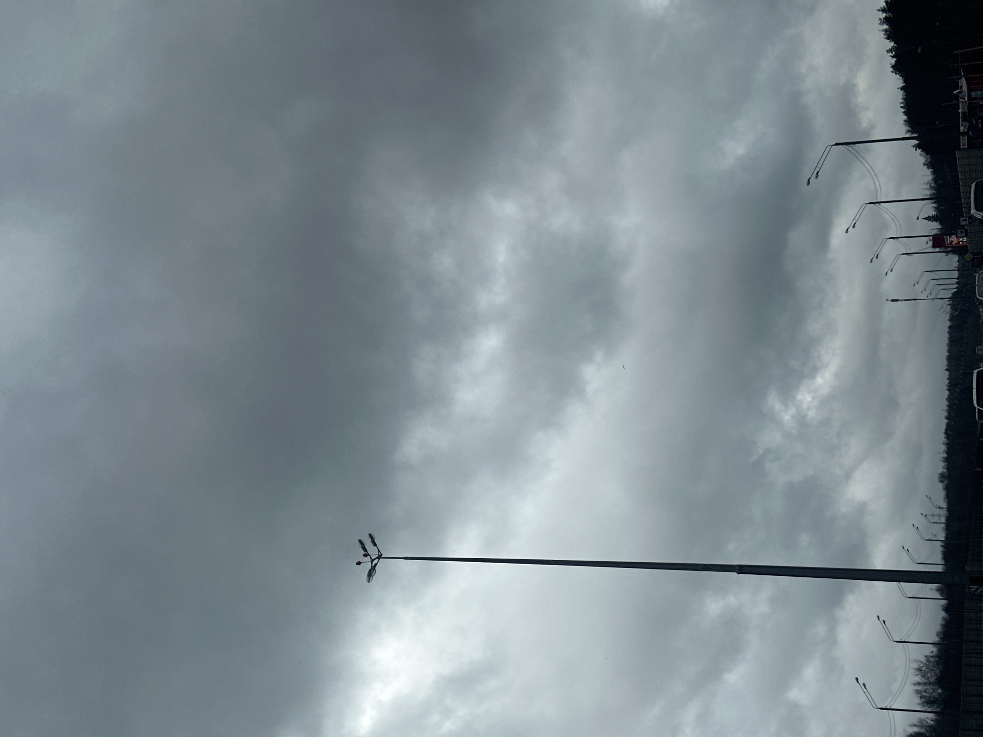 The image depicts a cloudy sky over Velednikovo, Russia. A tall lamppost with multiple lights extends upward from the bottom center of the frame. In the background to the right, a line of trees and other lampposts is visible, suggesting a road or highway. There is also parking along this line. The general atmosphere is overcast and gray.