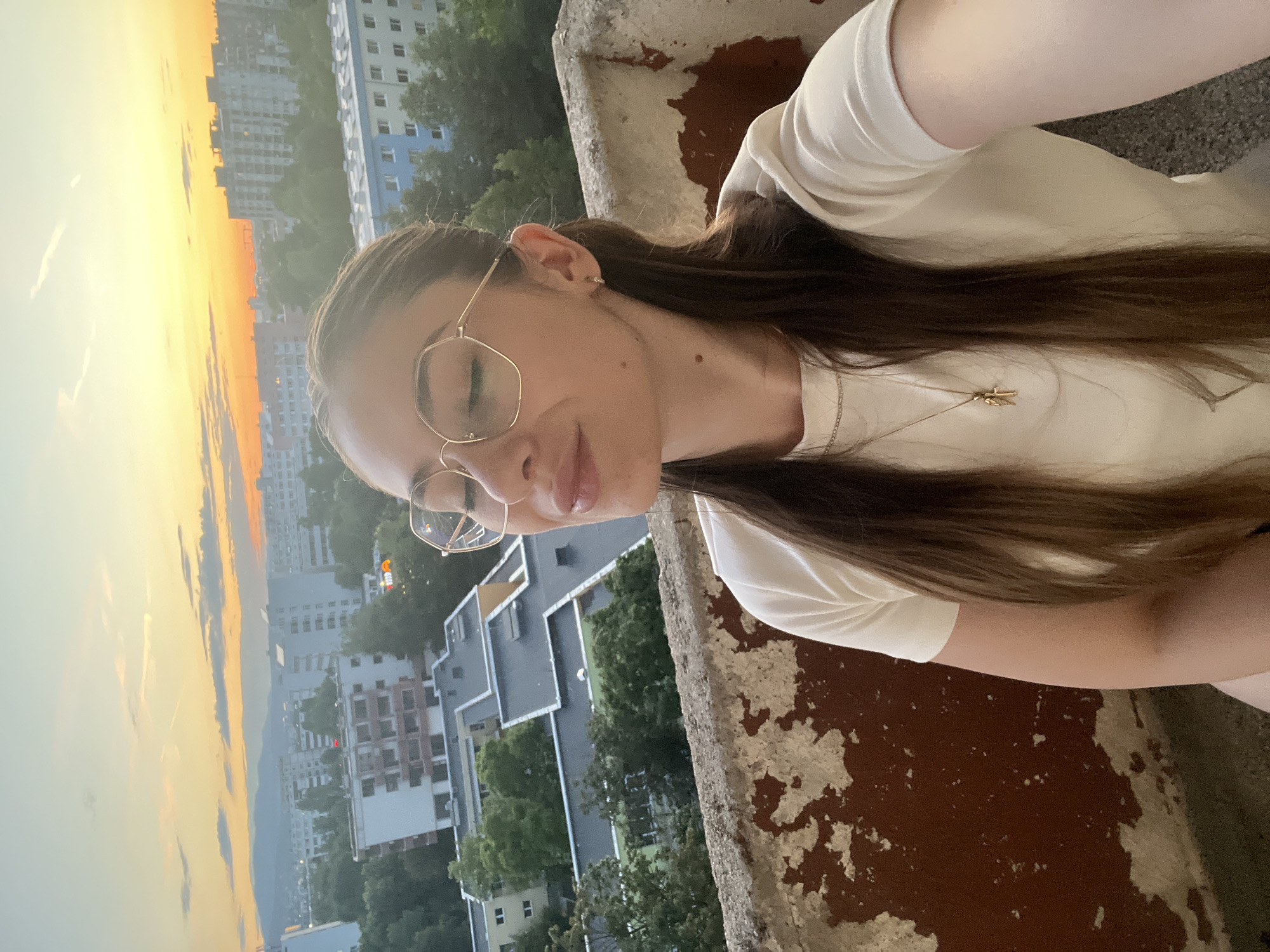 A young woman with long, dark brown hair and glasses is taking a selfie on a balcony overlooking a city at sunset. The woman is wearing a white t-shirt and a silver necklace with a gold pendant. The sky is a vibrant orange and yellow, with clouds streaking across it. In the background, there are several apartment buildings and trees. The balcony railing is made of concrete and has peeling paint.
