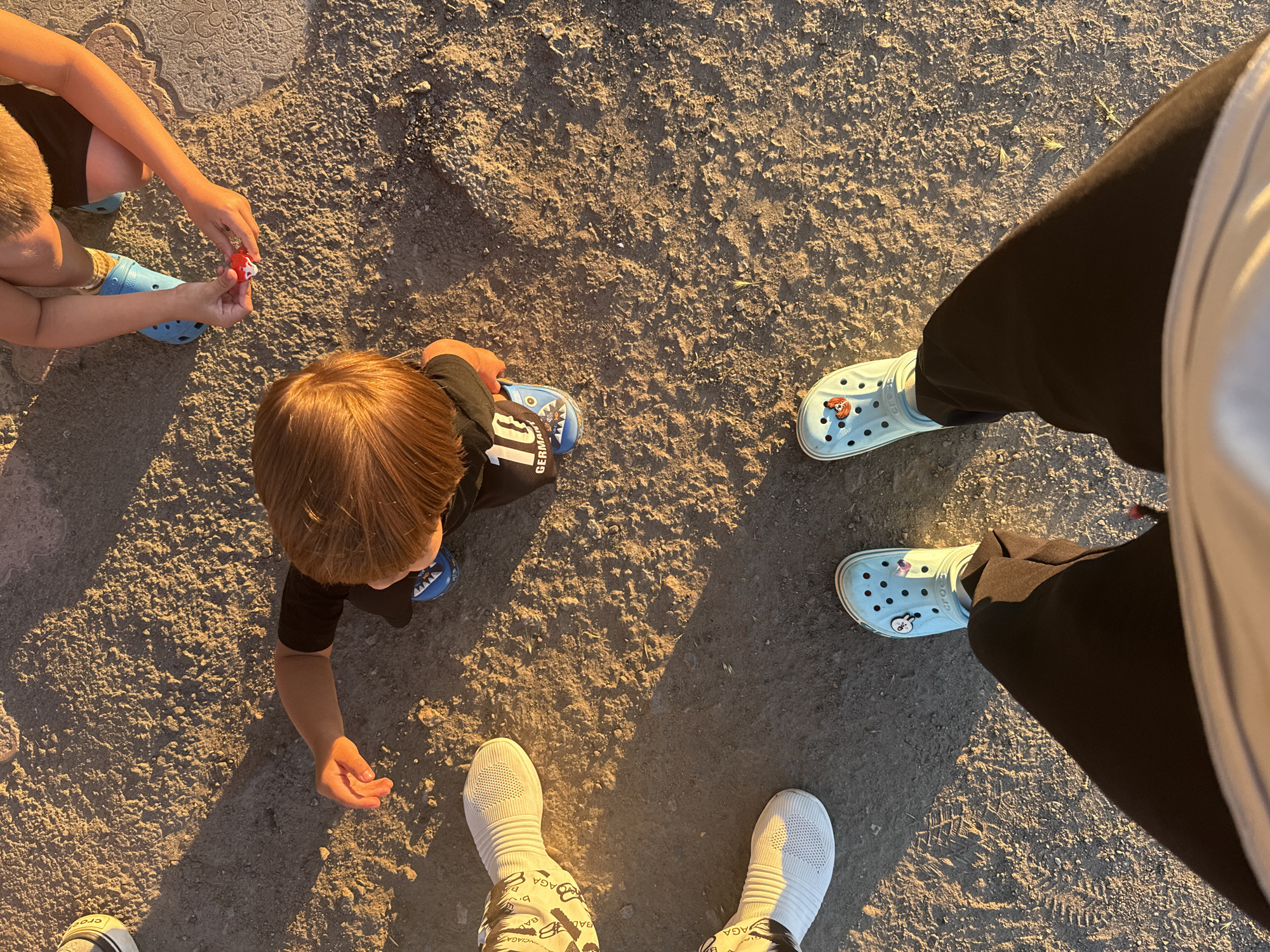 The image shows an aerial view of several pairs of feet and lower legs on a sandy, outdoor ground, possibly in Dnipro, Ukraine. The sunlight casts long shadows, indicating it is either early morning or late afternoon.
In the center, a young child with reddish-brown hair is wearing a black t-shirt with "10 GERMANY" written on it and blue Crocs with shark designs. The child's hand is reaching out towards a small red object.
To the left, another child's legs are visible, wearing blue Crocs.
Further to the left, two pairs of feet are visible. One pair is wearing white socks with a black and white pattern. The other pair is wearing white mesh sneakers.
In the foreground, a person is wearing black pants and light blue Crocs adorned with small charms. The person's legs and feet occupy the bottom portion of the frame.
The ground appears to be a mix of sand and small stones, with some tire tracks visible. The overall environment suggests an outdoor, potentially recreational area.
