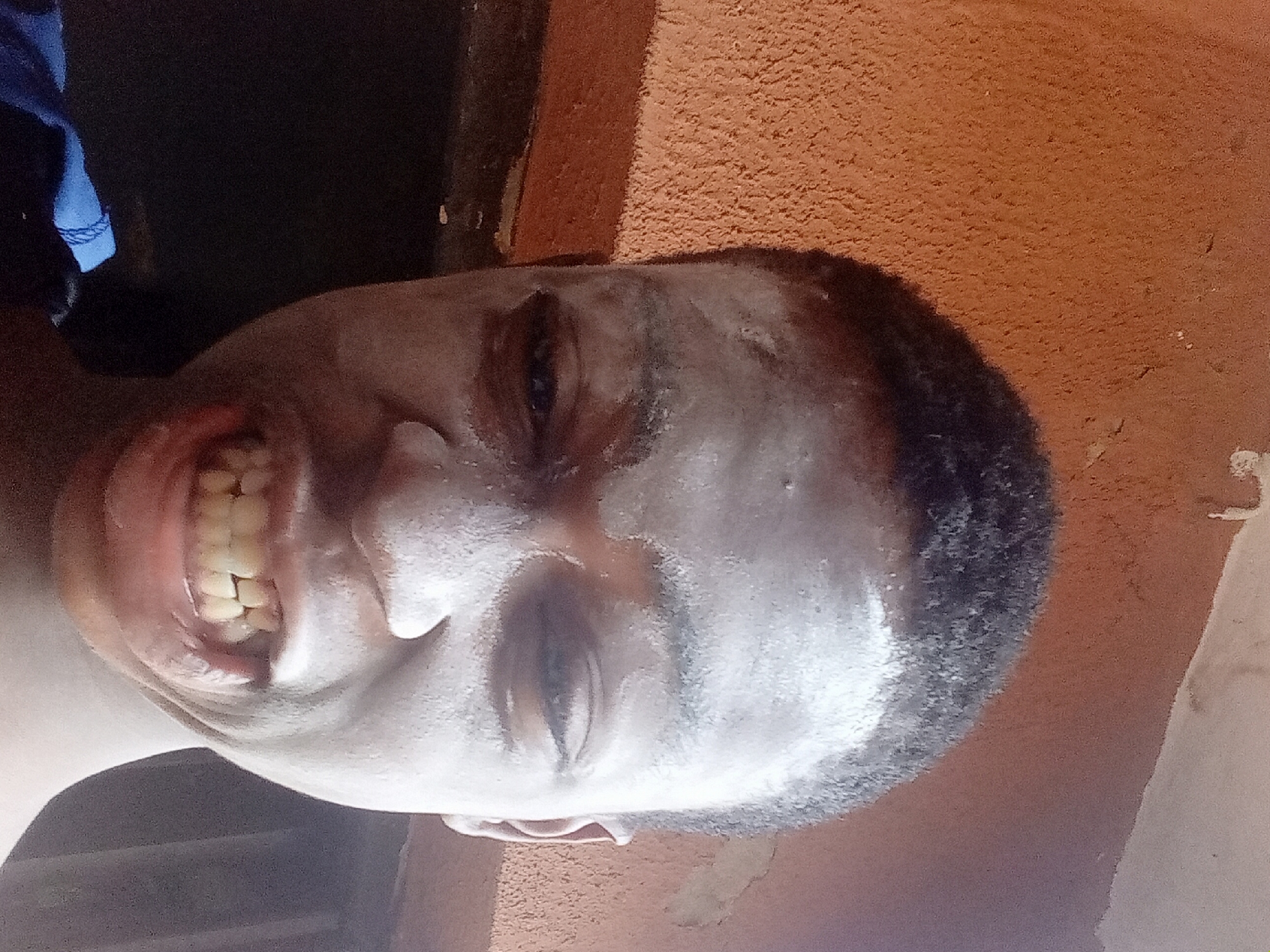 The image is a close-up of a person's face with a light-colored cream or substance applied to their face. The person is smiling, revealing their teeth. The background includes a dark, possibly interior space, and a textured, reddish-brown surface, likely a wall. The image was taken in Nkpor, Nigeria.