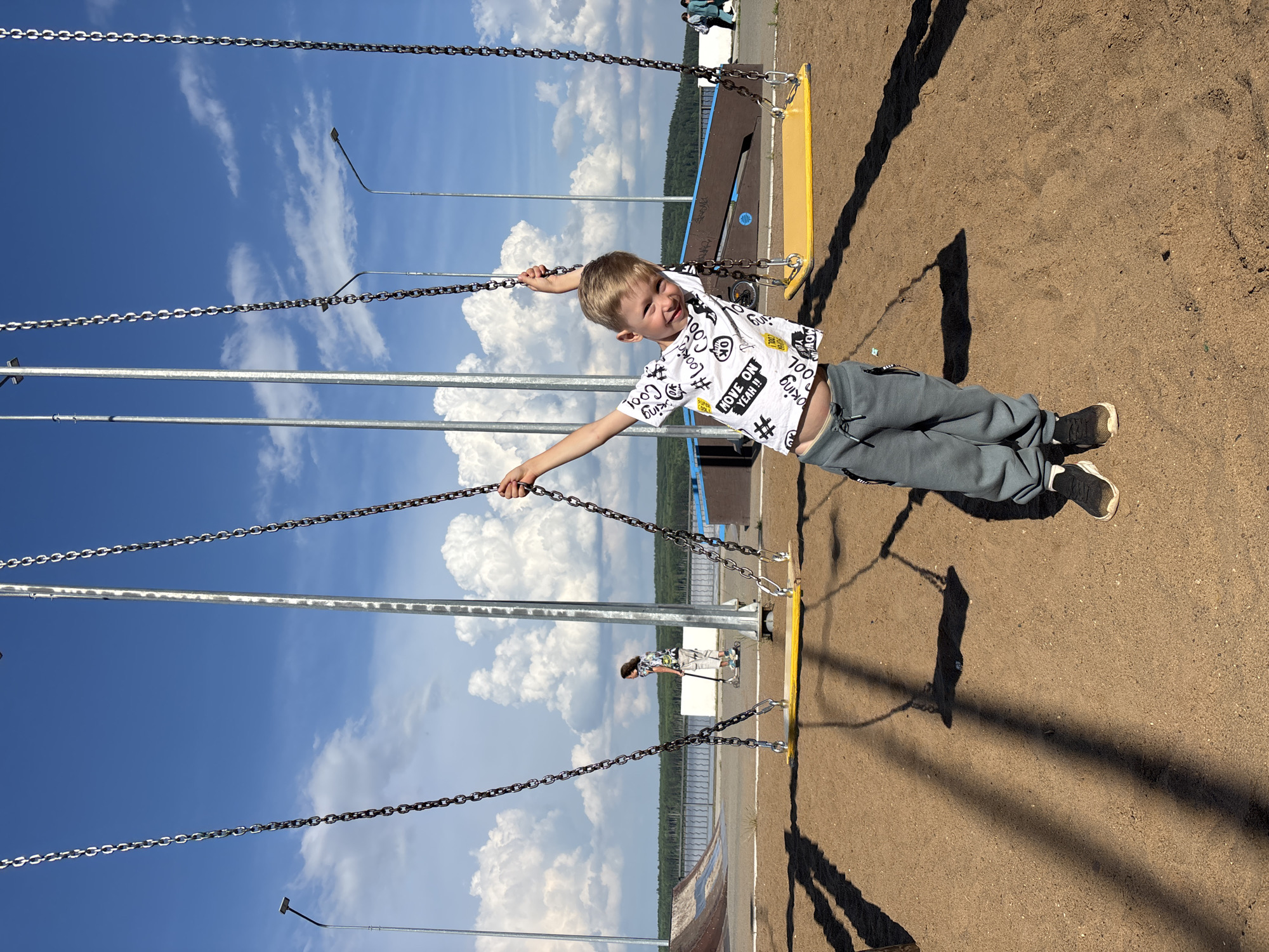 An outdoor shot captures a young boy with blond hair, wearing a white t-shirt with black text and graphics, and grey sweatpants, standing in a sandbox beneath a swing set. The boy is smiling and holding onto the swing chains with outstretched arms. In the background, a girl with dark hair is seen riding a scooter. Further in the background, there is a body of water, trees, a skate park with ramps, and streetlights under a partly cloudy blue sky. This scene is located at the "Sestra" waterfront in Chaikovsky, Russia.