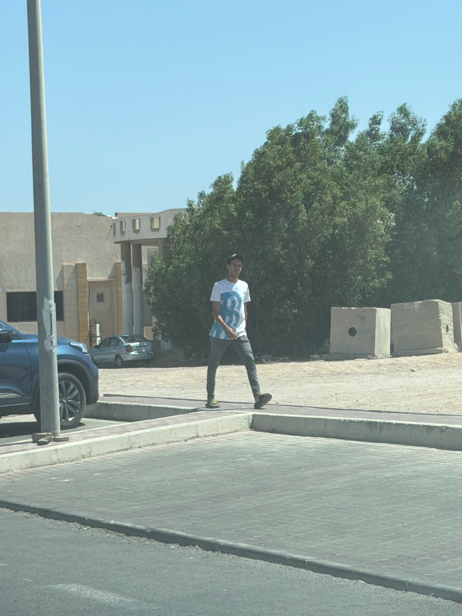 A young man wearing a white t-shirt with a blue geometric design and dark pants is walking on a sidewalk next to a street. He is facing towards the left of the frame. In the background, there is a light-colored building with rectangular openings, lush green trees, and two large concrete blocks. A blue car is parked on the left side of the image, and a light-colored sedan is visible further in the background. The scene appears to be an outdoor urban environment.