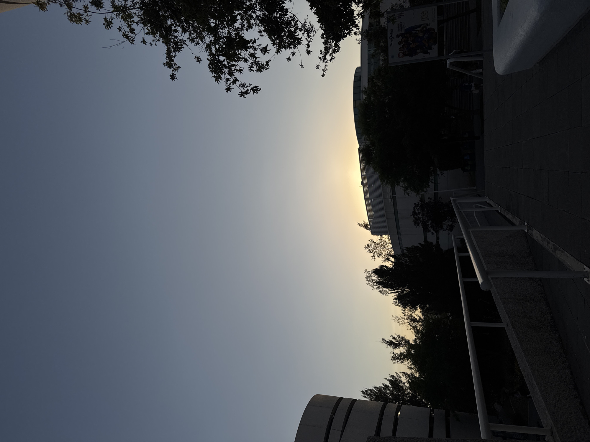 The image depicts an outdoor scene at dusk, likely in Puebla, Mexico, given the architectural style and vegetation. The sky transitions from a deep blue to a lighter, golden hue near the horizon, suggesting the sun is setting or rising. Silhouetted trees and architectural structures frame the sky. There are buildings, concrete walkways, and metal railings on one side of the frame, and the top left corner shows tree branches with dense foliage. The lighting is dim and creates stark contrasts between light and shadow.