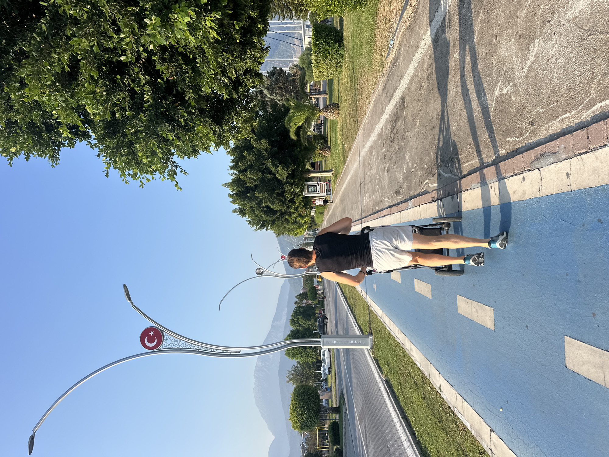A man wearing a black tank top and white shorts is pushing a stroller along a blue-painted bicycle path. The path has white dashed lines marking it. To the left of the path is a road with cars and a sidewalk. In the background, mountains are visible. There are streetlights with a Turkish flag emblem. To the right of the path is a grassy area with trees. The scene appears to be in Fethiye, Turkey, as indicated by the streetlights and the overall landscape. The sun is shining, casting shadows on the path.