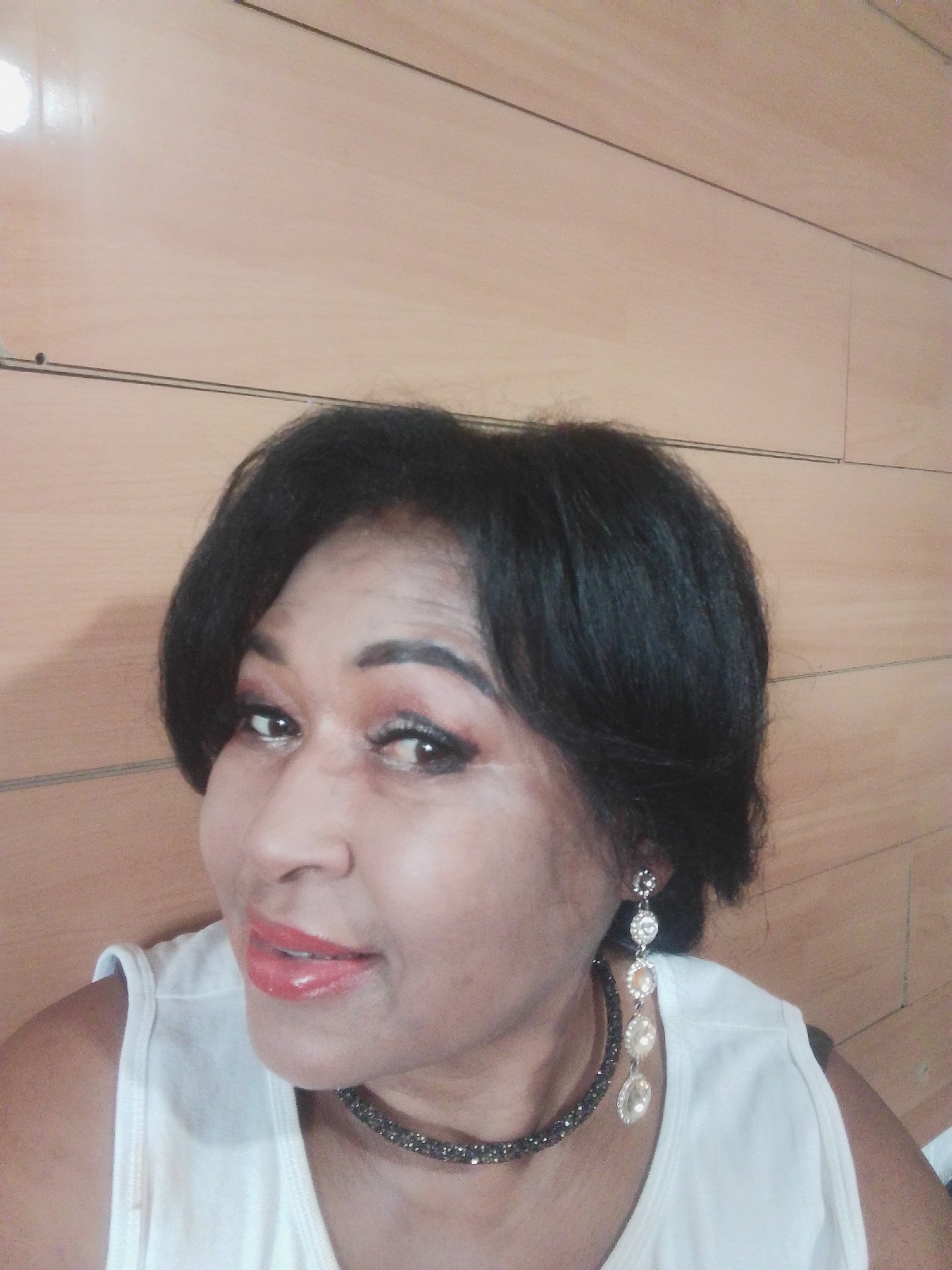 A medium shot selfie shows a Black woman with dark, layered hair, wearing a white sleeveless top, a sparkling dark choker necklace, and a dangling earring with multiple clear stones. She has on makeup, including red lipstick and eyeshadow. The background is a wood-paneled wall. The image provides no information about the city of Kuntuu, Kyrgyzstan.
