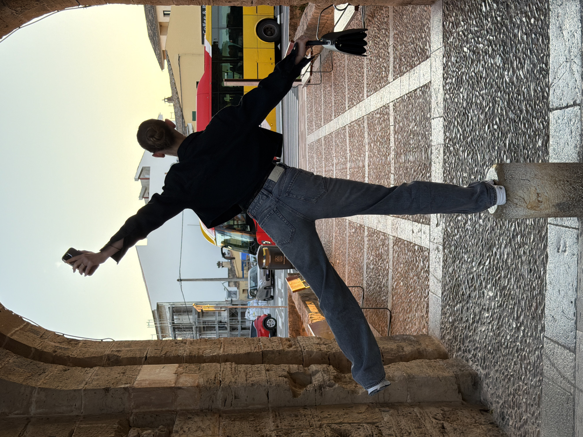 The image shows a person standing with their legs spread, one foot on a stone pillar, under an archway. The person is wearing a black jacket and gray jeans. They are holding a cell phone in one hand and an umbrella in the other, with their arms outstretched to either side. The archway is made of rough, aged stone. Beyond the archway, there is a street scene including a yellow and red bus, cars, and buildings in Alcúdia, Spain. The ground is paved with stone tiles. The sky is a pale yellow, suggesting it is either early morning or late evening.