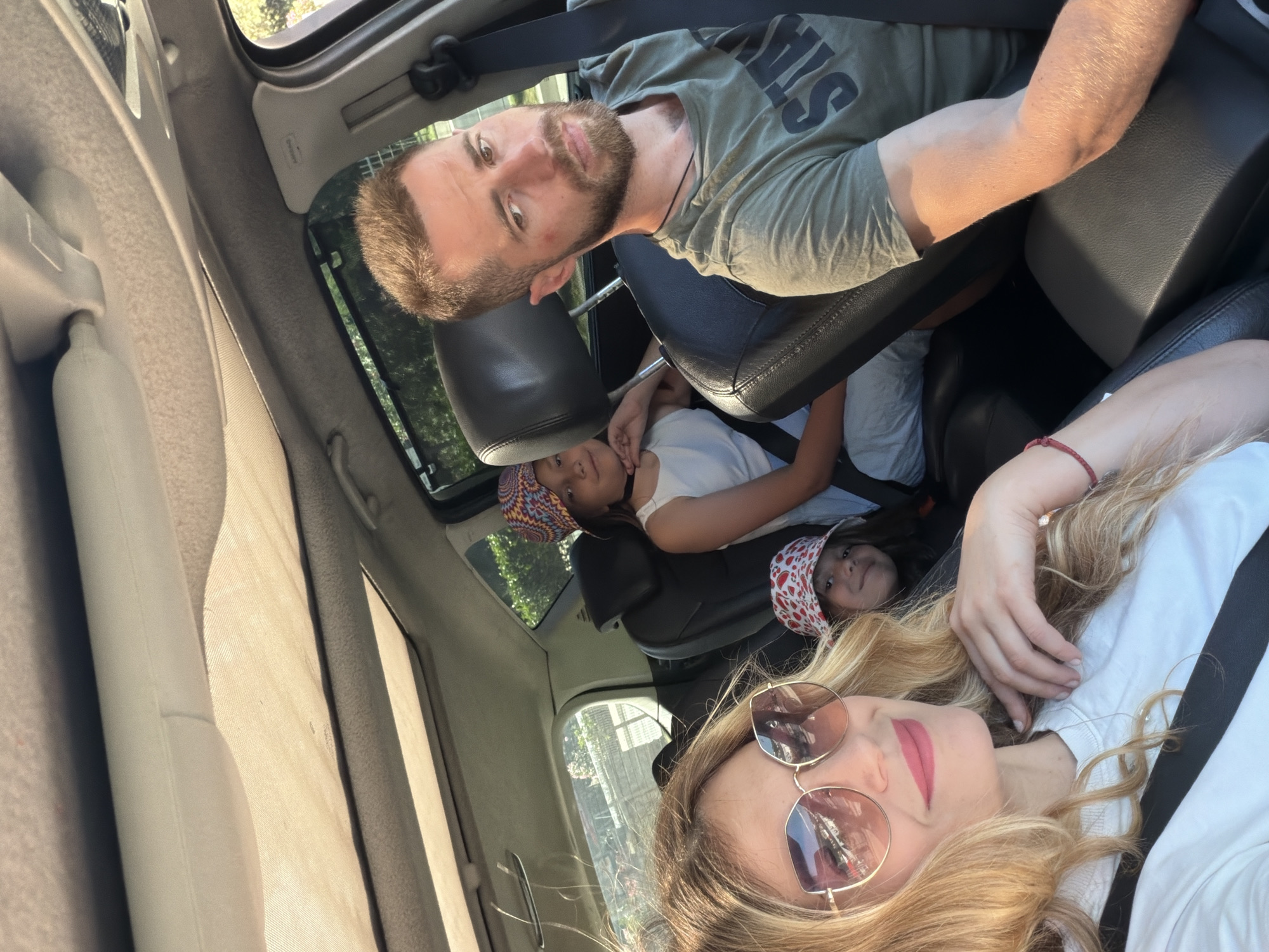 A family of four is depicted inside a vehicle, likely in the city of Budva, Montenegro, during daylight hours. A blonde-haired woman, wearing sunglasses and a white t-shirt, occupies the front passenger seat, taking a selfie of the group. She has pink lipstick and her left hand, adorned with a red bracelet, is visible. A dark-haired man with a beard, wearing a green t-shirt with partial text "STA" visible, is in the driver's seat, looking towards the camera. Both adults are wearing seatbelts. In the rear passenger seats, two children are visible. The child behind the driver, a girl, wears a multi-colored patterned bucket hat and a white top, and is looking towards the camera with her chin resting on her hand. The child behind the front passenger, also a girl, wears a white bucket hat with red strawberry patterns; only part of her face is visible as she smiles towards the camera. The car's interior features dark seats and a light-colored headliner with an open sunroof, through which sunlight streams. Black mesh sunshades cover parts of the rear windows. Outside the vehicle, green foliage, trees, and parts of light-colored architecture are visible. An "Airbag" label is discernible on the passenger-side A-pillar.