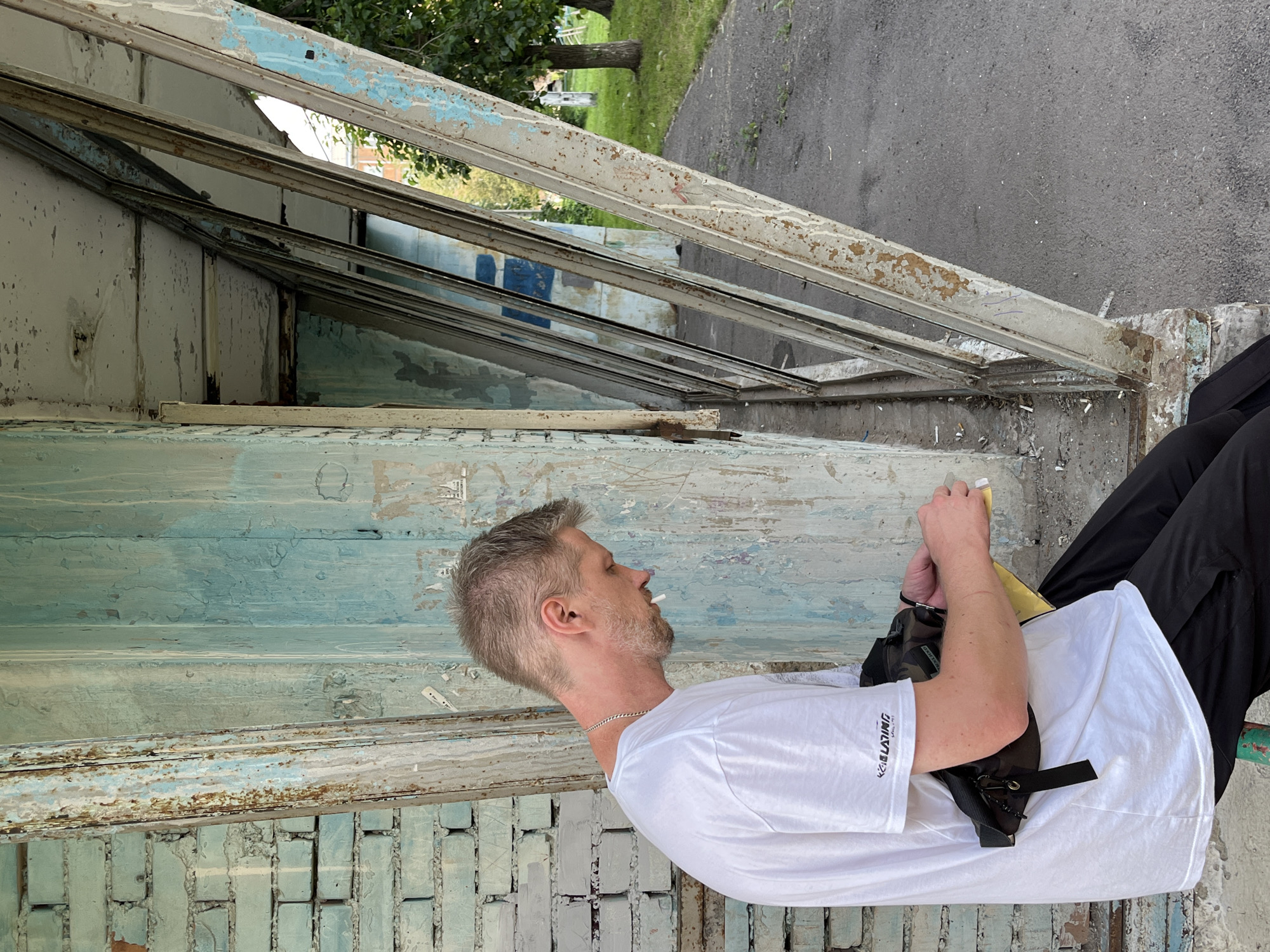 A man with graying hair and a beard is seen from the side, wearing a white t-shirt and black pants. He has a fanny pack worn across his chest. The man is holding a cigarette and appears to be smoking. He is sitting outdoors in an urban environment, with a brick wall to his left and a structure with metal beams and peeling paint to his right. The ground is paved with asphalt, and there is grass and trees in the background. The scene depicts a moment of rest or contemplation in a somewhat dilapidated outdoor setting. This location is likely a bus stop or a similar public waiting area, given the presence of a bench and the overall utilitarian architecture.