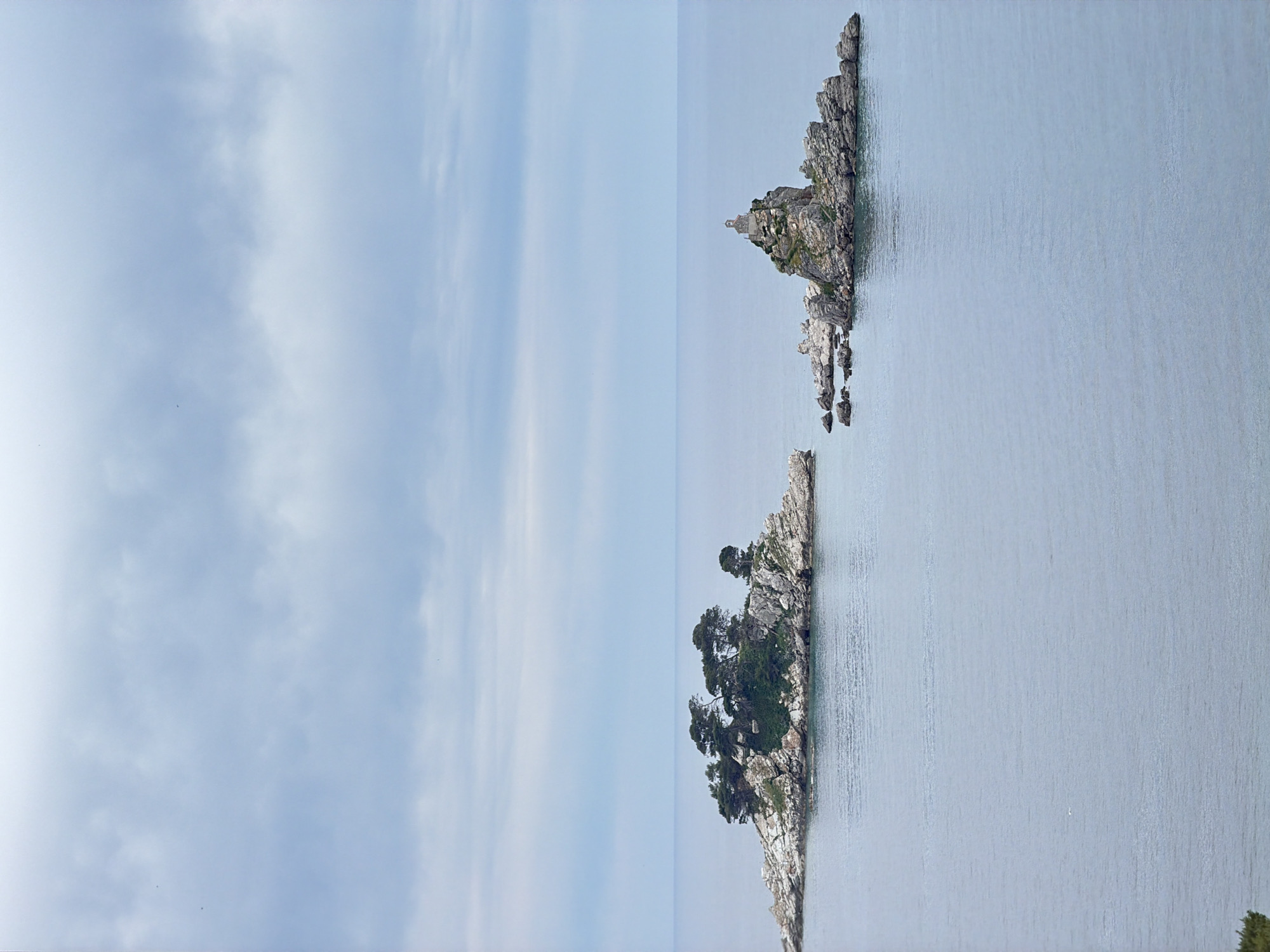 The image captures a serene seascape featuring two rocky islets in the Adriatic Sea, specifically near Petrovac na Moru, Montenegro. The sky, covering the left portion of the image, is light blue with scattered clouds. To the right, the calm water reflects the sky's color. The islets are located towards the bottom-right portion of the image. The islet on the upper-right features a church or monument on its peak, while the larger islet below is covered with trees. The overall atmosphere is tranquil, emphasizing the natural beauty of the Montenegrin coast.