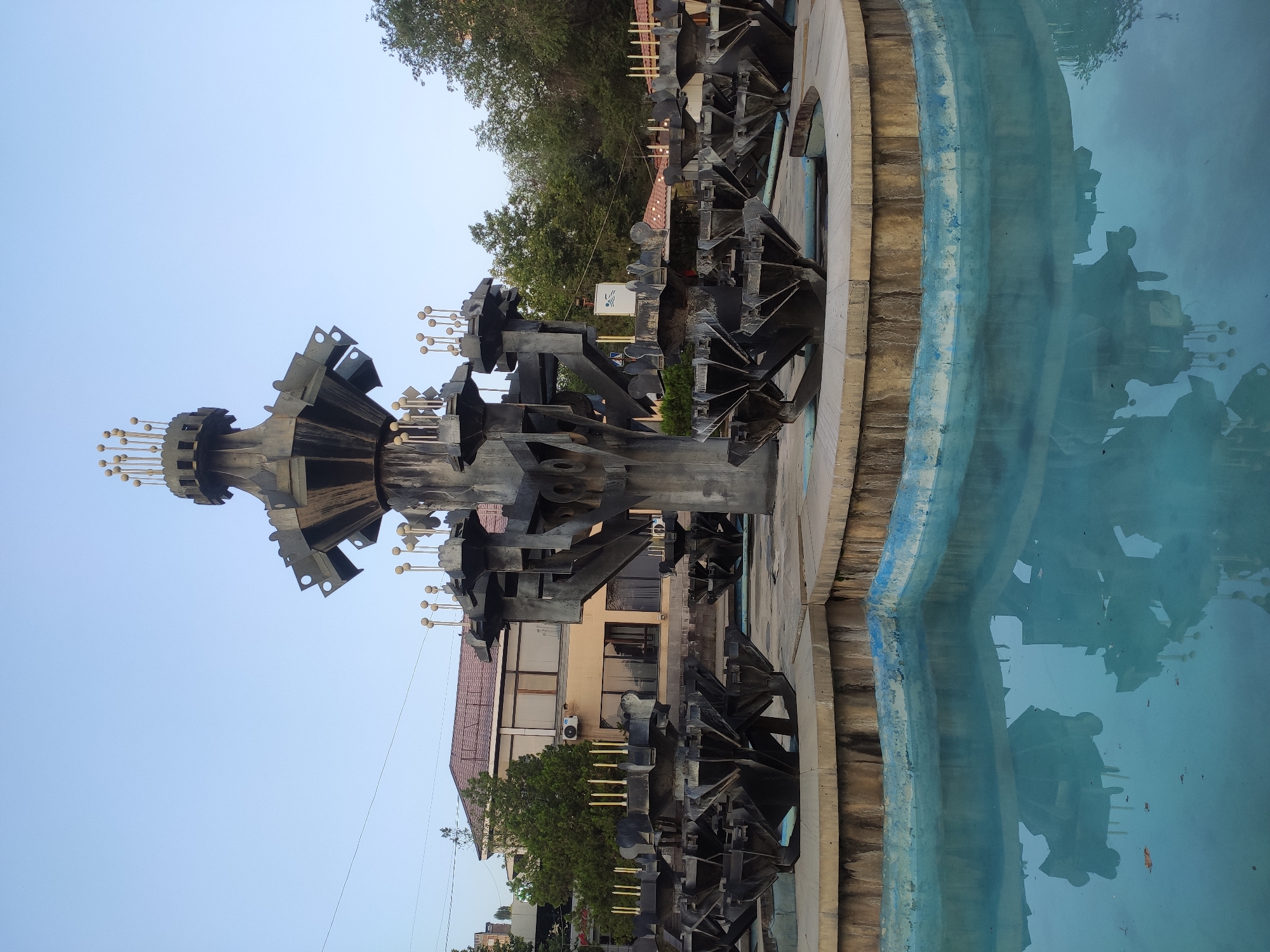 The image shows a fountain with a central sculptural element, located in Yerevan, Armenia. The fountain has multiple tiers of dark, angular metal structures, each topped with small, spherical ornaments. The water in the basin is a clear blue and reflects the fountain's structure and the surrounding environment. In the background, there are buildings with brown roofs and beige facades, along with trees and a clear blue sky. The scene appears to be outdoors and during daylight hours.