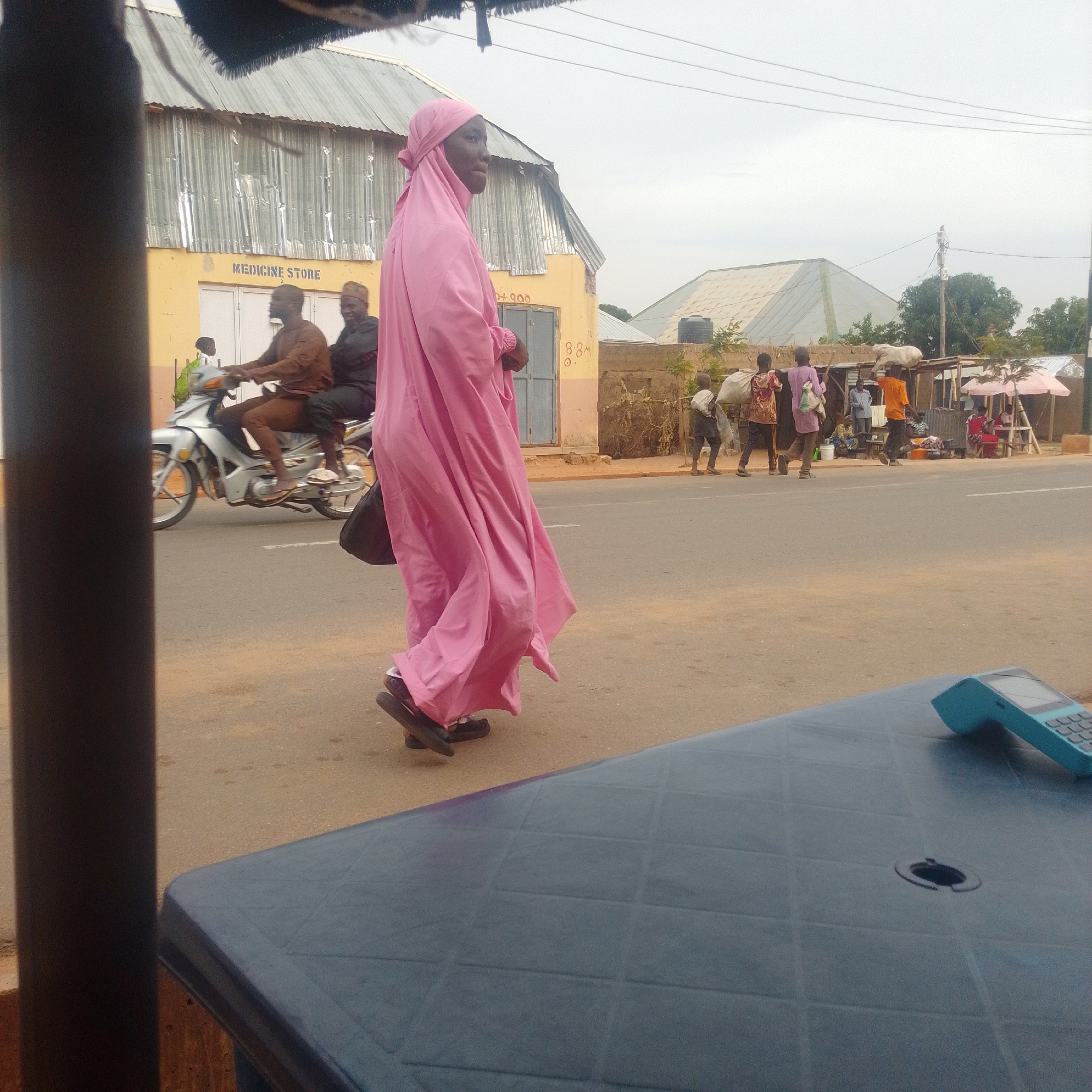 In Gombe, Nigeria, a medium shot captures a street scene with a focus on a person walking away from the camera, clad in a bright pink hijab and flowing garment. The individual wears dark sandals. To the left, a man and a woman ride a silver motorcycle, with the man driving and the woman seated behind him. The building behind them, painted yellow, displays the sign "MEDICINE STORE" in orange lettering. Further in the background, several other individuals are visible, some carrying large bags on their heads, suggesting market activity. The environment includes a paved road, dirt shoulders, and various structures, including shops with awnings. The sky is overcast. In the foreground, the corner of a dark blue table with a textured surface is visible, with a blue credit card reader resting on it.