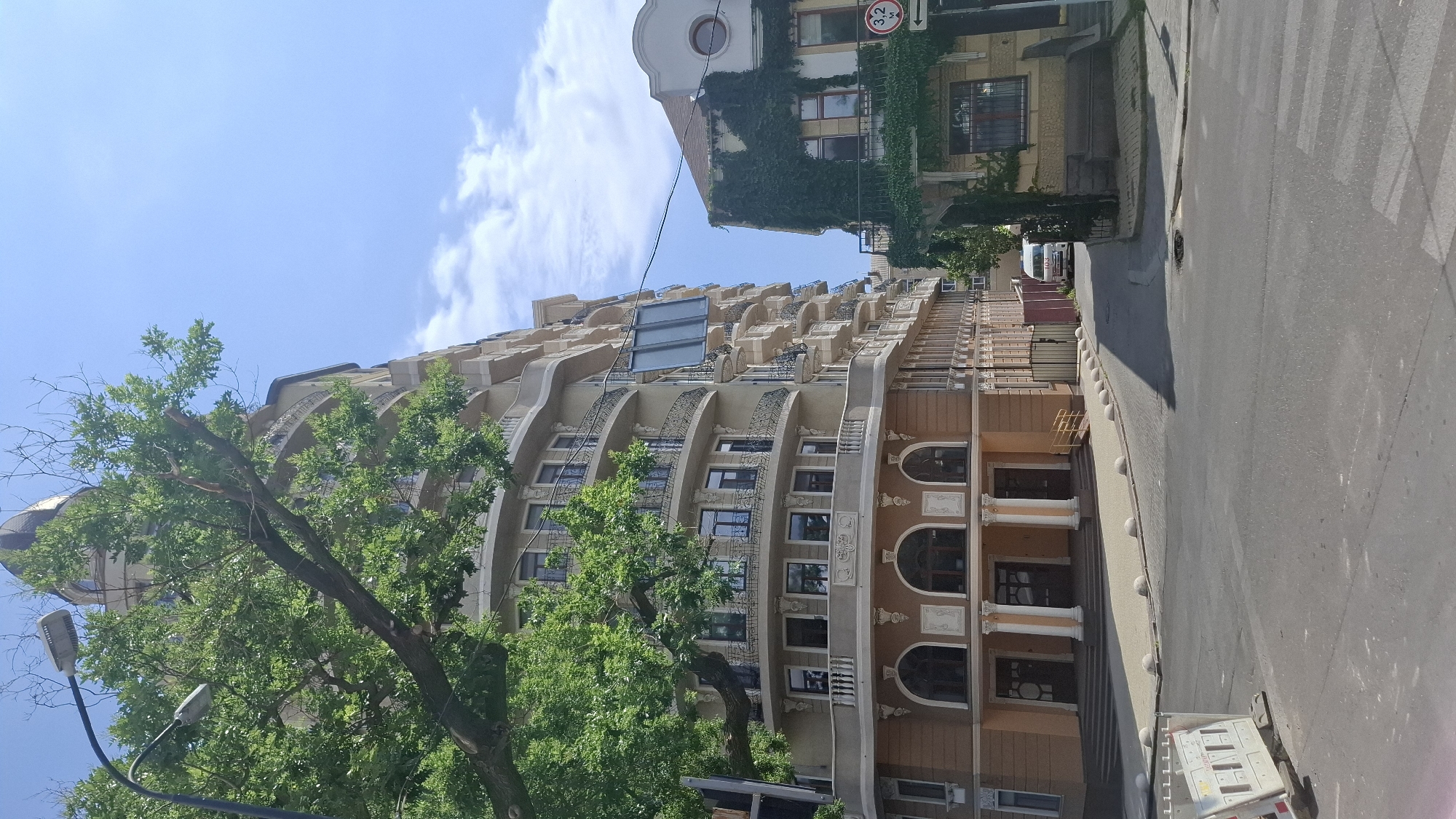 An outdoor, eye-level, medium shot captures a street scene in front of two buildings. The building on the left is a large, light-colored, multi-story structure with a curved facade and numerous balconies. It features arched windows on the lower level and a rounded dome-like structure at the top. A large, leafy green tree partially obscures the upper portion of this building. The building on the right is smaller and appears older, covered in dense green ivy. It has a single circular window on its upper level and a balcony with a railing. A street sign indicating a height restriction of 3.2 meters and a directional arrow is visible on the right side of this building. The street in the foreground is paved, with a pedestrian crossing marked. A white utility box is present in the lower left corner. In the distance, a white van is parked on the street between the two buildings. The sky is blue with scattered white clouds.