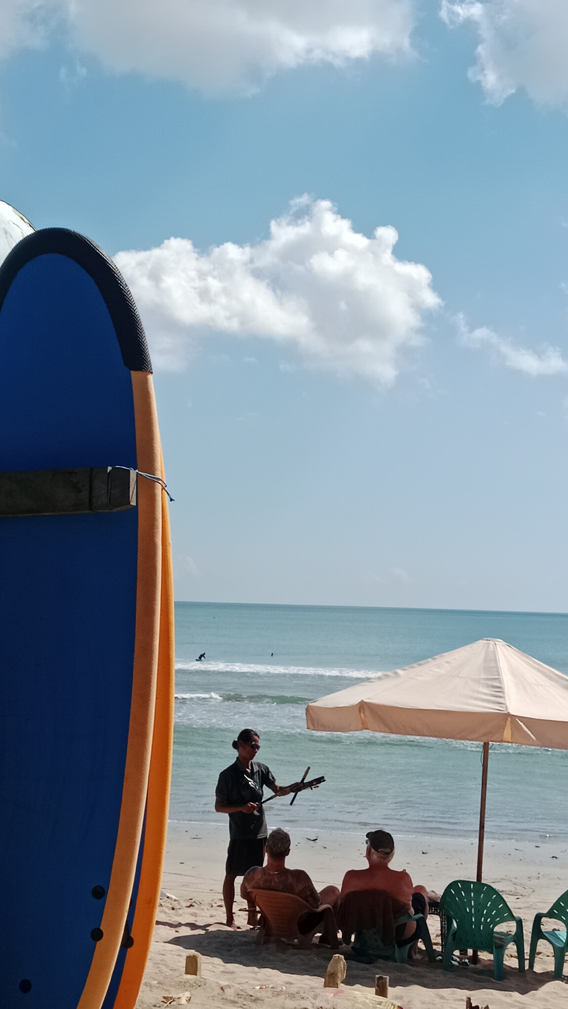 A close-up of a large blue and orange surfboard is in the foreground on the left, obscuring part of the scene. To the right, on a sandy beach in Kuta, Indonesia, a person with dark hair pulled back stands facing away from the camera, holding a small, dark, stick-like object with several extensions. They are wearing a dark t-shirt and dark shorts. To their right, two people are seated in lounge chairs. The person on the left is facing away from the camera and the person on the right is partially facing the camera. Both are wearing hats. Behind them, an umbrella with a cream-colored canopy is open. In the background, the turquoise ocean stretches to the horizon under a bright blue sky with scattered white clouds. There are small waves breaking on the shore, and at least two surfers can be seen in the water.