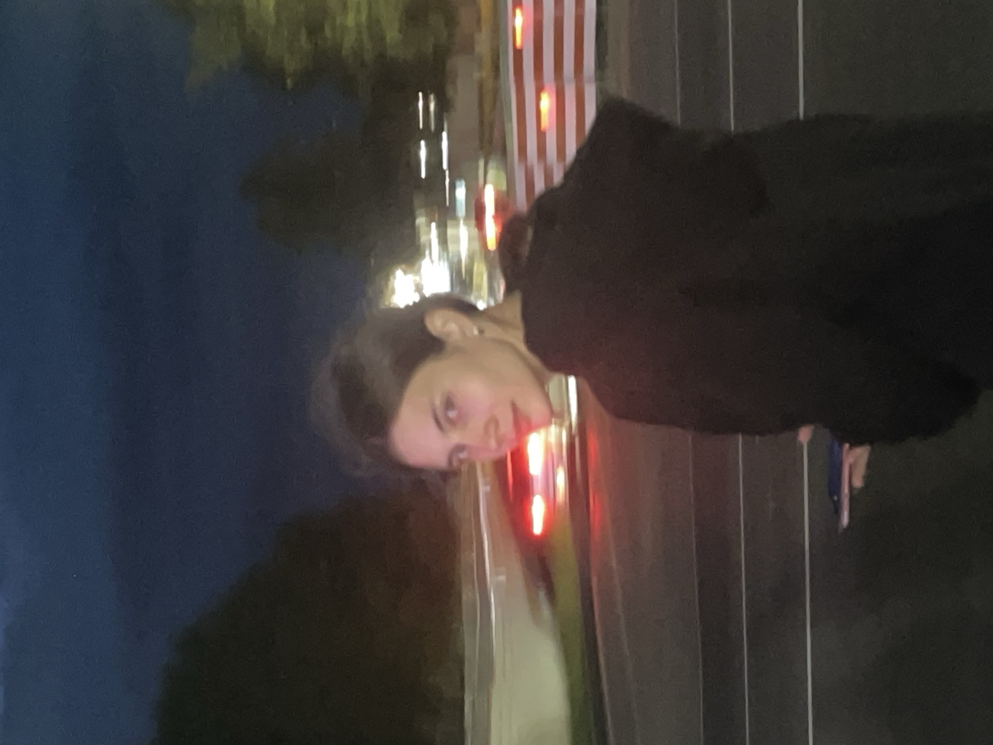 The image shows a woman with dark hair pulled back, wearing a dark hooded jacket, looking over her right shoulder at the camera. She is holding a small dark object in her right hand, possibly a phone. The background is blurred, suggesting motion, and depicts a street scene at dusk or night. Red taillights from a vehicle are visible on the left side of the image, and horizontal red and white striped barriers are present in the mid-ground. The sky is dark blue. The location is identified as Votkinsk, Russia, suggesting an urban or suburban street setting within that city.