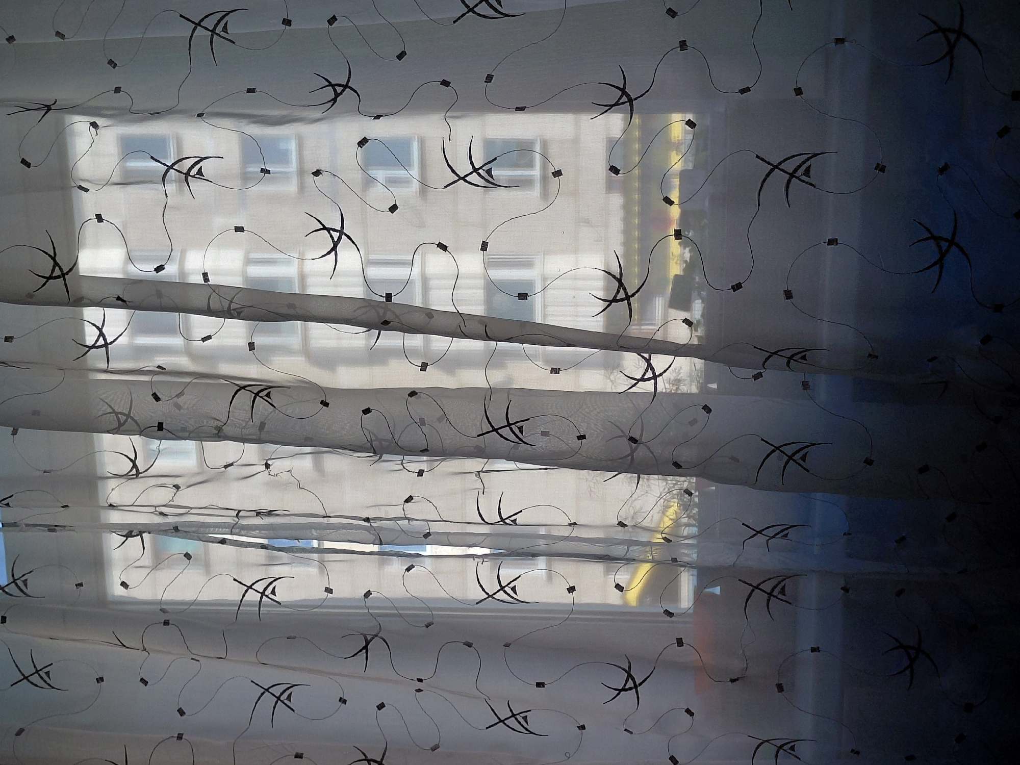 The image displays an indoor perspective looking outwards through a sheer, light-colored curtain. The translucent curtain, likely a Roman blind or a pleated panel, is adorned with an abstract pattern of dark, stylized curvilinear lines and small square motifs. These decorative elements are repeated across the fabric, which appears to be layered or folded horizontally, creating variations in transparency and shadow.
Beyond the curtain and window, the blurred outline of a light-colored, multi-story building is visible. This building features multiple rectangular windows, suggesting an urban or residential environment, typical for a city like Nadym, Russia. Bright daylight streams in from outside, indicating it is daytime with clear weather. The light illuminates the exterior building and filters through the curtain, highlighting its texture and design while creating a soft glow.
No people are present in the image, nor is any specific activity depicted; it is a static scene observing the view from within. There is no visible text. The overall impression is one of calm and domesticity, with the decorative curtain providing a gentle separation from the outside world.