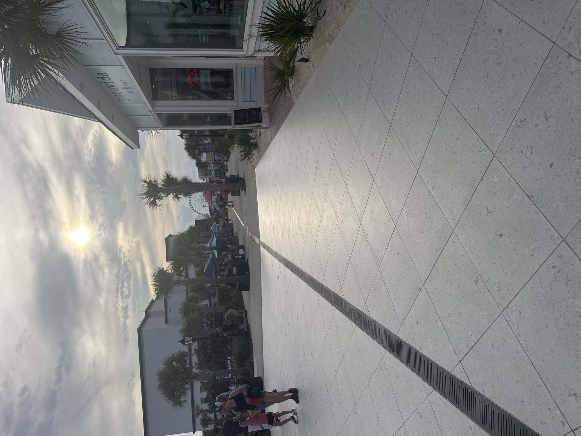 The image depicts a scene at the Myrtle Beach Boardwalk and Promenade in Myrtle Beach, South Carolina, United States of America. The viewpoint is from a paved walkway constructed with large, square, light-colored tiles, a dark metal drain running diagonally across the foreground. To the right, a building with light blue siding and large glass windows is visible, with a sign that reads "puravida" and "LIVE FREE". In front of the building, there are some small green bushes planted in sand.
Further down the promenade, palm trees line the walkway, with a Ferris wheel visible in the distance. Several people are walking along the promenade. In the mid-ground on the left, a woman in a black tank top and denim shorts is walking with a young girl in a pink tank top and black shorts. The sky is overcast with the sun peeking through, creating a slightly hazy atmosphere. The overall impression is of a pedestrian area, likely a tourist destination, on a day with dynamic weather conditions.