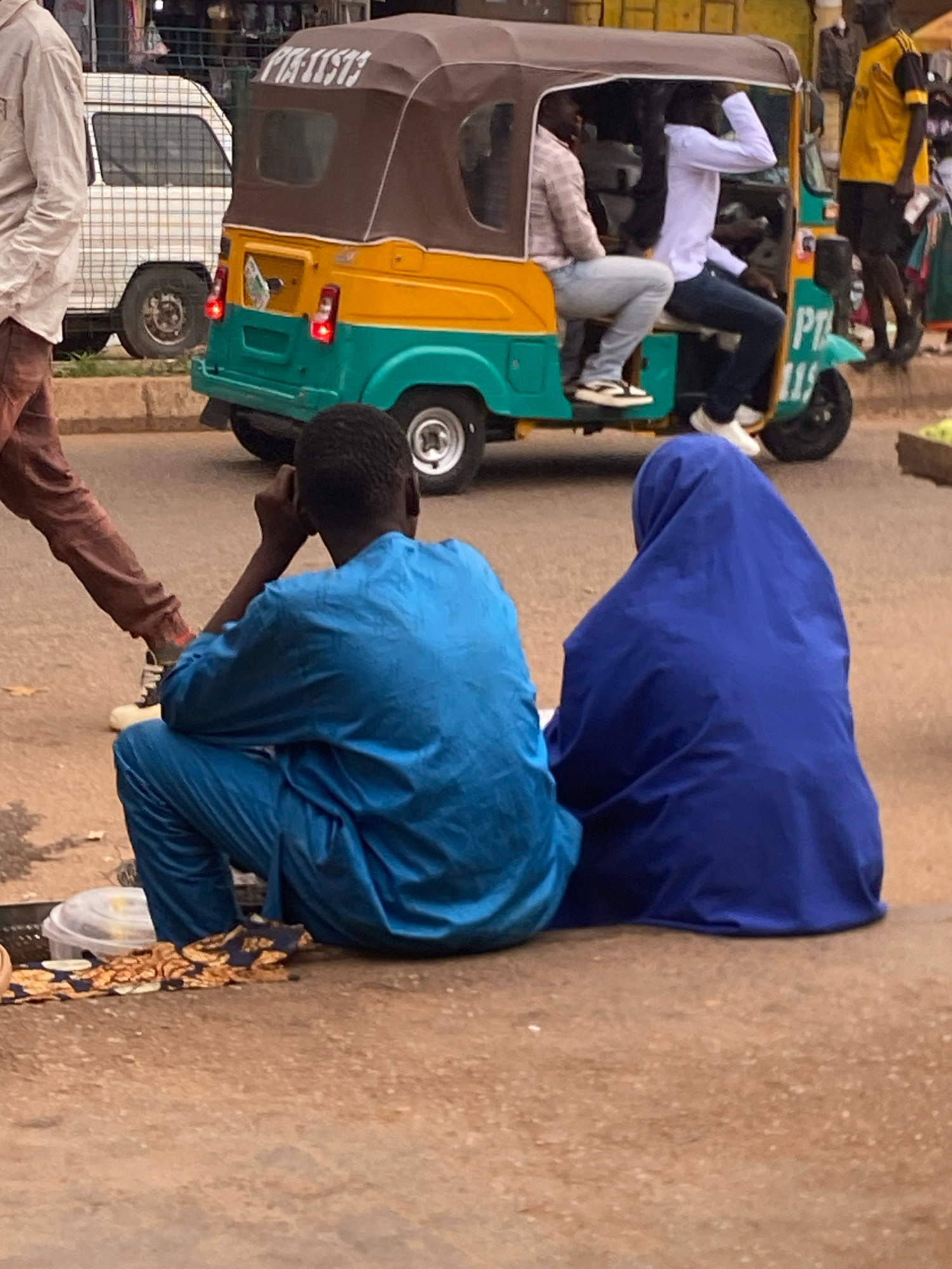 In Bukuru, Nigeria, two individuals are seated on a dusty, unpaved road. A person on the left, facing away from the camera, is dressed in a blue tunic and trousers. Next to them, a person wearing a dark blue hijab is also seated, facing away from the camera. In the background, a three-wheeled auto-rickshaw, painted yellow and teal, is stopped on the road. The auto-rickshaw has a brown canopy and appears to be carrying passengers. A white van with a checkered pattern on its side is parked to the left of the auto-rickshaw. The scene suggests a typical street environment in an urban area.