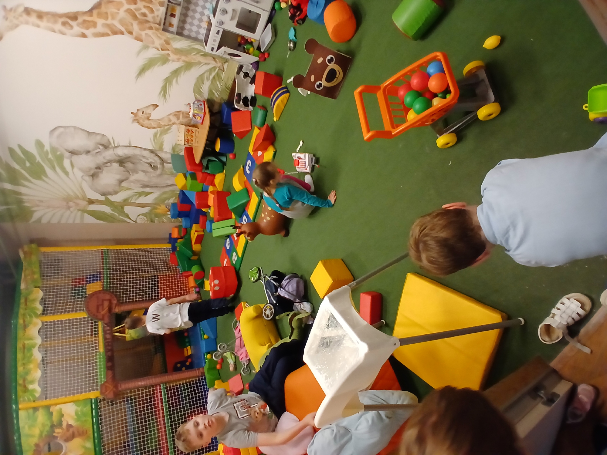 The image depicts an indoor children's play area, likely a soft play center, with a green carpeted floor. Several children are present. In the foreground, a young boy in a light blue shirt is partially visible from the back, looking down. To his right, a white sandal is on the floor. Further back, a boy in a gray t-shirt and dark pants is seated on an orange cushion. To his left, a white high chair is prominent. A girl in a teal shirt and light pants is crouched down, playing with a toy. Scattered throughout the space are numerous colorful soft play blocks, a small orange toy shopping cart filled with colored balls, a deer-shaped toy, a toy kitchen, and other miscellaneous toys. In the background, a multi-level play structure with netting and jungle-themed decorations is visible. The walls are adorned with a mural featuring jungle animals, including an elephant and giraffes. The scene suggests a lively and active play environment. There is no information available to definitively place this location in Yekaterinburg, Russia.