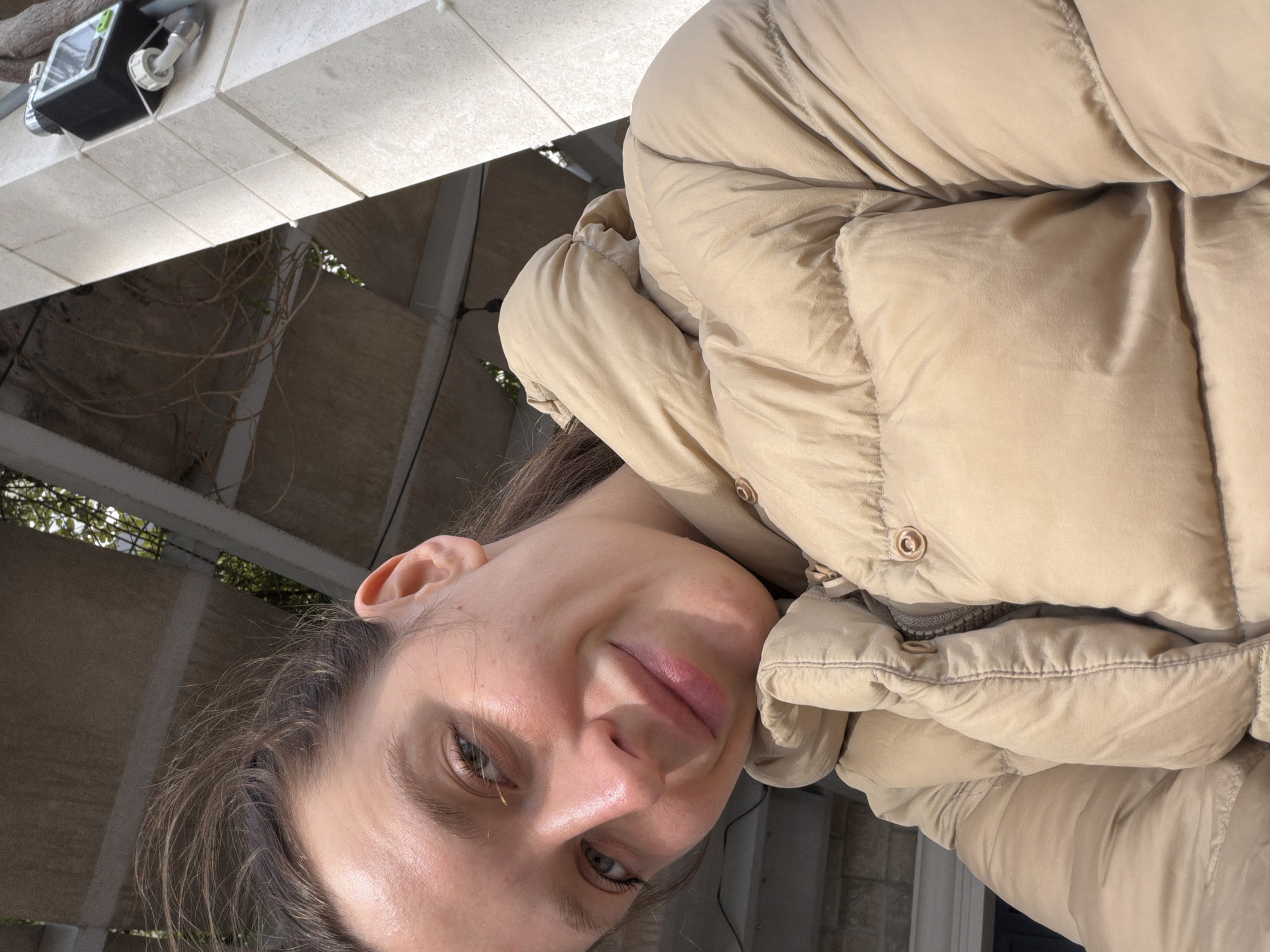 A woman wearing a tan puffer jacket is taking a selfie in Rafailovići, Montenegro. The location appears to be outdoors, possibly on a balcony or under a covered area, judging by the architectural elements in the background. The background shows a tiled surface and some vines. A small device with wires is attached to the surface above her. The focus is on the woman's face, her jacket, and the immediate surroundings.