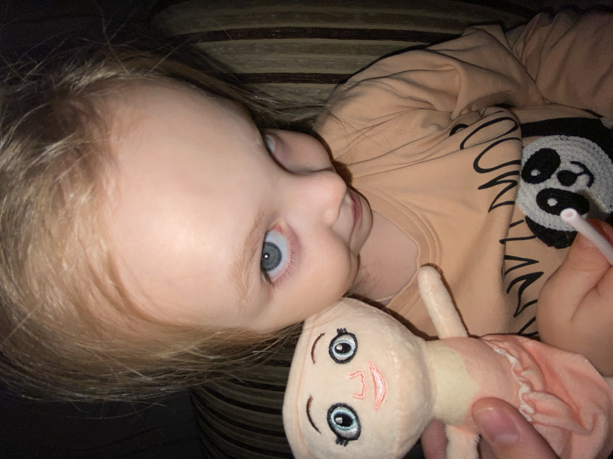 This media file presents a close-up, high-angle shot of a young child, likely a toddler, with fair hair and striking blue eyes, looking upwards directly at the camera. The child is positioned indoors, possibly resting on a striped surface, such as a couch or bed, indicated by the dark and light striped fabric in the background.
The child is wearing a light peach or beige long-sleeved shirt, which features a partially visible black graphic or text on the front and a white and black crocheted panda design. A person's hand is visible on the right side of the frame, holding a small plush doll with an embroidered smiling face and blue eyes, dressed in pink. The hand also holds a white straw, positioned near the doll.
The child's expression appears calm and engaged, suggesting a moment of quiet play or interaction. The lighting is soft, consistent with an indoor setting, rather than bright daylight. The overall scene conveys a tender and intimate moment. The location context for this image is Votkinsk, Russia.
