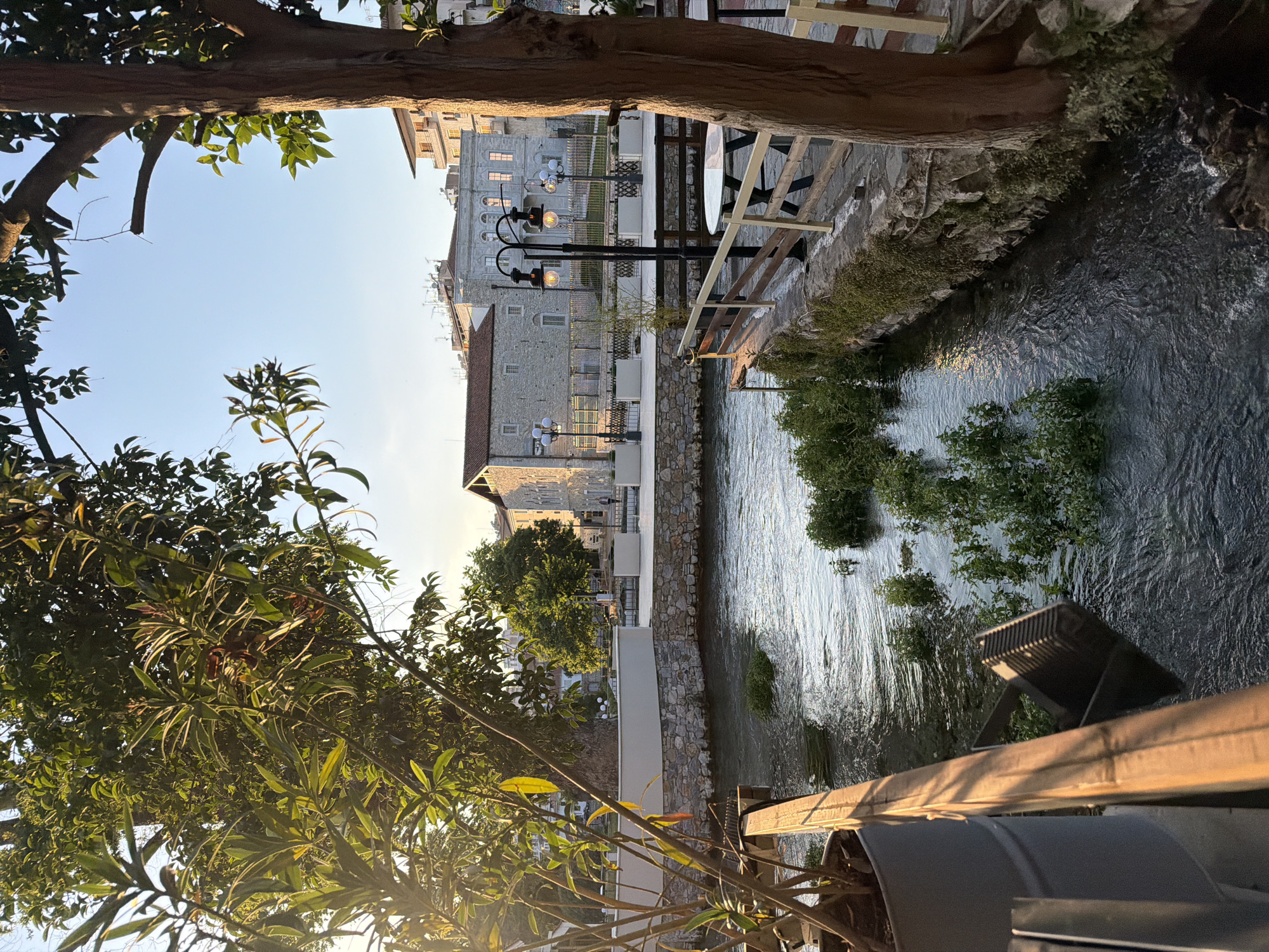 The image presents a view of the city of Drama in Greece. The scene is framed by trees and their foliage in the foreground, partially obscuring the overall view. A river runs through the center of the frame, reflecting the ambient light. The river banks are lined with stone walls and vegetation. Beyond the river, buildings with light-colored facades and traditional architectural elements are visible. Street lamps are positioned along the river and near the buildings, indicating urban development. Tables and chairs can be seen suggesting a restaurant or cafe seating area adjacent to the river. The sky is clear and bright, suggesting daylight conditions.