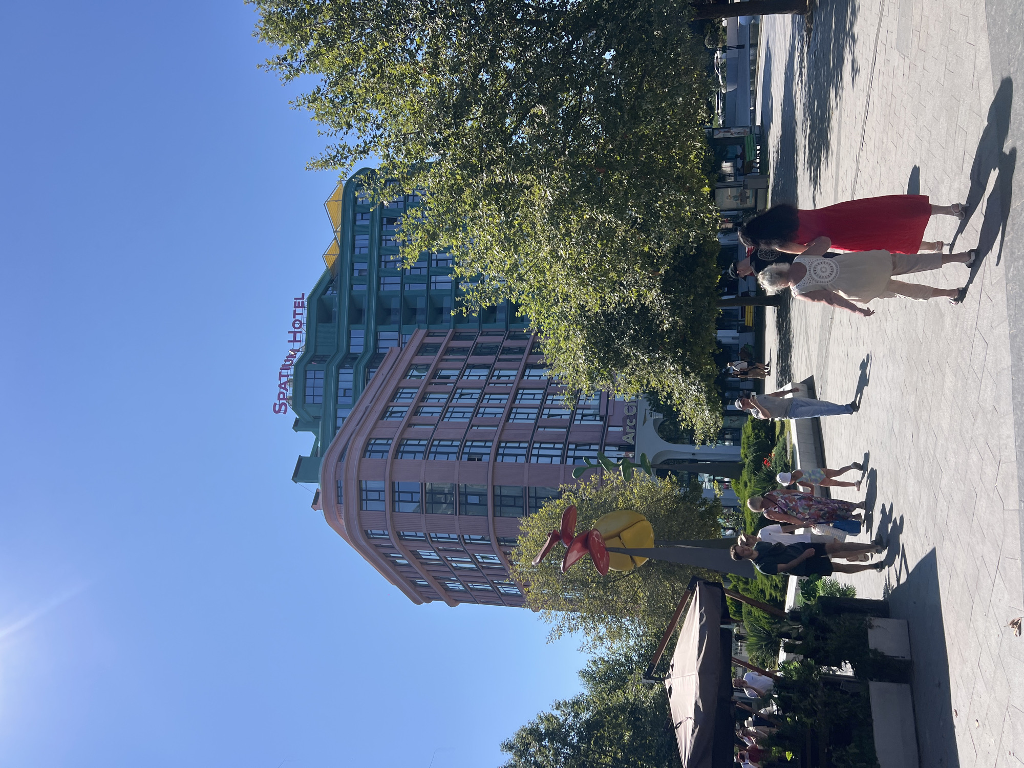The image depicts a sunny outdoor scene in Odesa, Ukraine, likely in a pedestrian area. In the background, a modern hotel with a distinct pink and green facade stands prominently. The hotel is topped with red lettering that reads "SPARATIUM HOTEL". To the left of the hotel, a large, abstract yellow and red sculpture is visible amidst lush green trees. In the foreground, several people are walking on a paved walkway. A group of individuals, including a man in a dark t-shirt and shorts, a woman in a floral dress, and a young child in a swimsuit and sunhat, are walking from left to right. Further to the right, two women are walking away from the camera, one in a red dress and the other in a cream-colored outfit. Outdoor seating areas with umbrellas are also present, suggesting a commercial or dining establishment. The clear blue sky and bright sunlight indicate a warm day.
