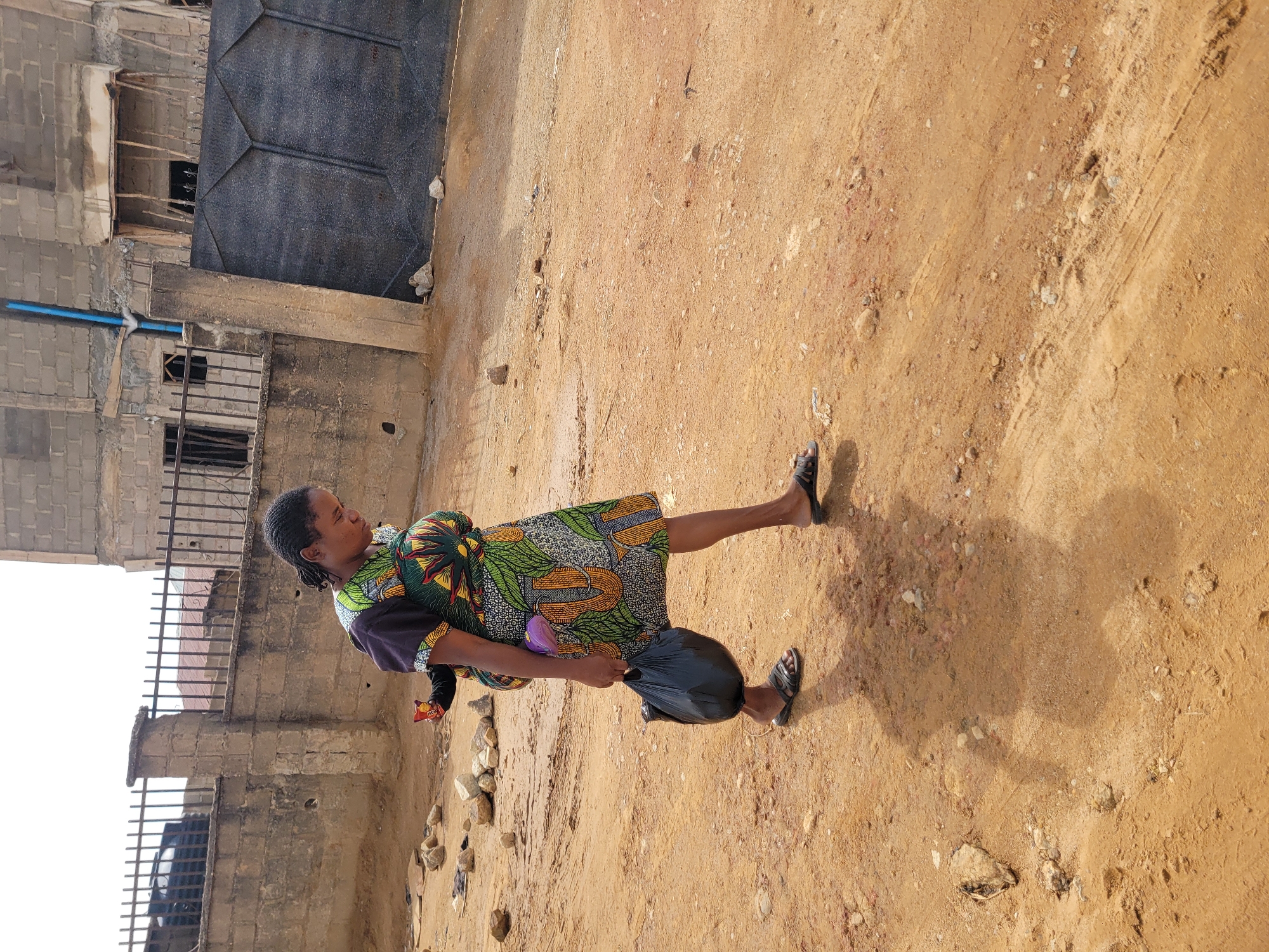 A woman stands on a dirt surface in Jos, Nigeria. She wears a patterned dress and sandals, holding a black plastic bag in her right hand. A child is secured on her back with a fabric wrap. Behind her are buildings constructed from cinder blocks. The ground is uneven and sandy.