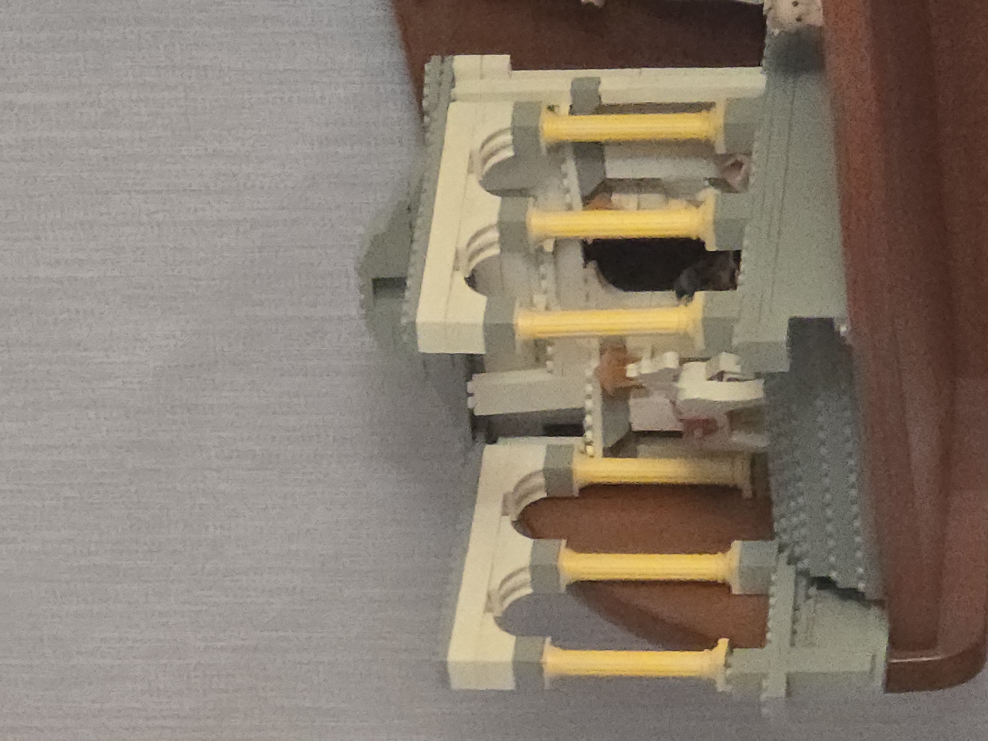 The image shows a close-up of a Lego architectural model resembling a classical building with columns and arches. The model is situated on a shelf with a textured, light gray wall behind it. The building features yellow columns, gray archways, and light beige upper structures. Stairs lead up to the entrance. The model appears to be assembled from standard Lego bricks. The lighting suggests an indoor setting.