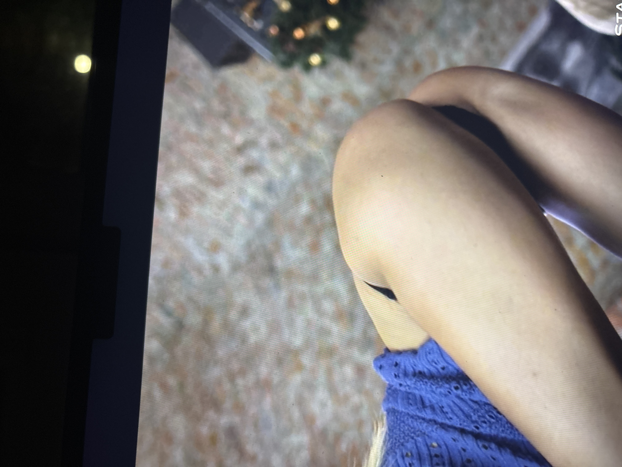 This image is a photograph of a digital screen, showing a partial view of a person in an indoor setting. The primary focus is on the person's bare legs, bent at the knees and positioned towards the center and right of the frame. The individual is wearing a blue, textured garment, possibly knitted or lacework, visible on the lower right. A distinct, dark, V-shaped mark or shadow is noticeable on the upper part of one leg.
The background of the screen content is softly blurred, revealing what appears to be a decorated Christmas tree with warm, glowing lights in the upper left section. A textured surface, resembling a light-colored carpet or rug, extends across the rest of the background.
The photograph itself captures reflections on the screen, including a bright, circular, yellowish light source on the dark, unlit portion of the screen's left edge. The image exhibits pixelation, indicative of a picture taken of a display rather than the original scene. In the top right corner of the displayed content, the vertically oriented text "STA" is partially visible. The overall impression is a cozy, relaxed indoor scene, likely during the evening or night, given the Christmas lights.