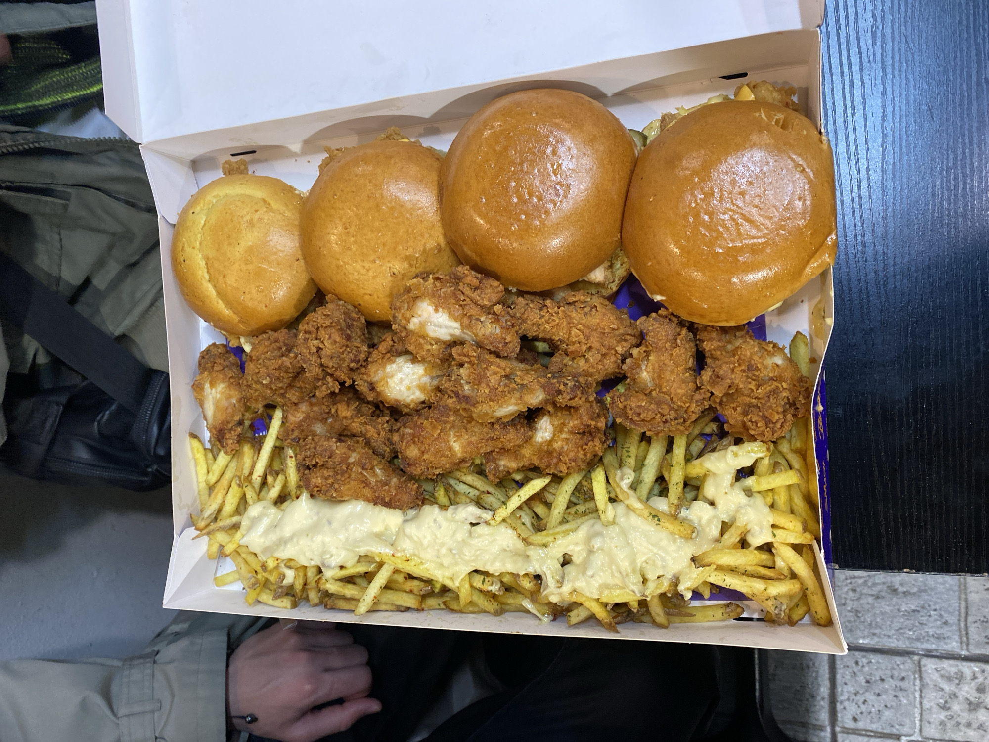 A top-down, medium shot shows an open cardboard box filled with food. The box contains four burger buns arranged at the top, below which is a layer of fried chicken pieces. Underneath the chicken, there is a bed of french fries topped with a white sauce. The box appears to be held by a person, as a hand and part of a jacket are visible on the left side of the frame. To the left of the box, there is a dark-colored bag. On the right side of the box, there is a surface with dark, vertical lines. This image was likely taken indoors, but the exact location in Paris, France, cannot be determined from the provided image.