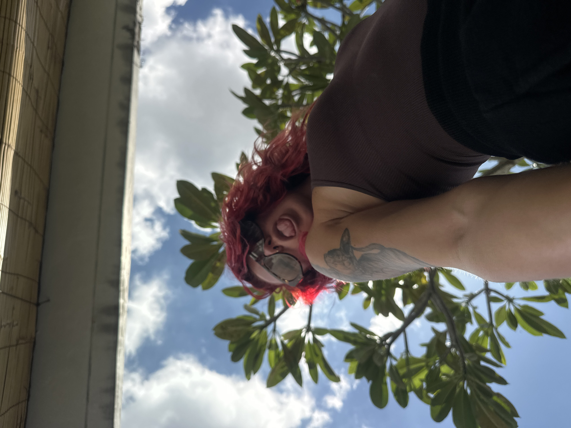 The image shows a low-angle view looking up at a person with red hair wearing sunglasses and a brown top, with a tattoo on their arm. They are sticking their tongue out. The background includes tree branches with green leaves, a blue sky with white clouds, and a building with a woven exterior. It is not possible to confirm the location in the city of North Kuta, Indonesia with the details provided in the image.