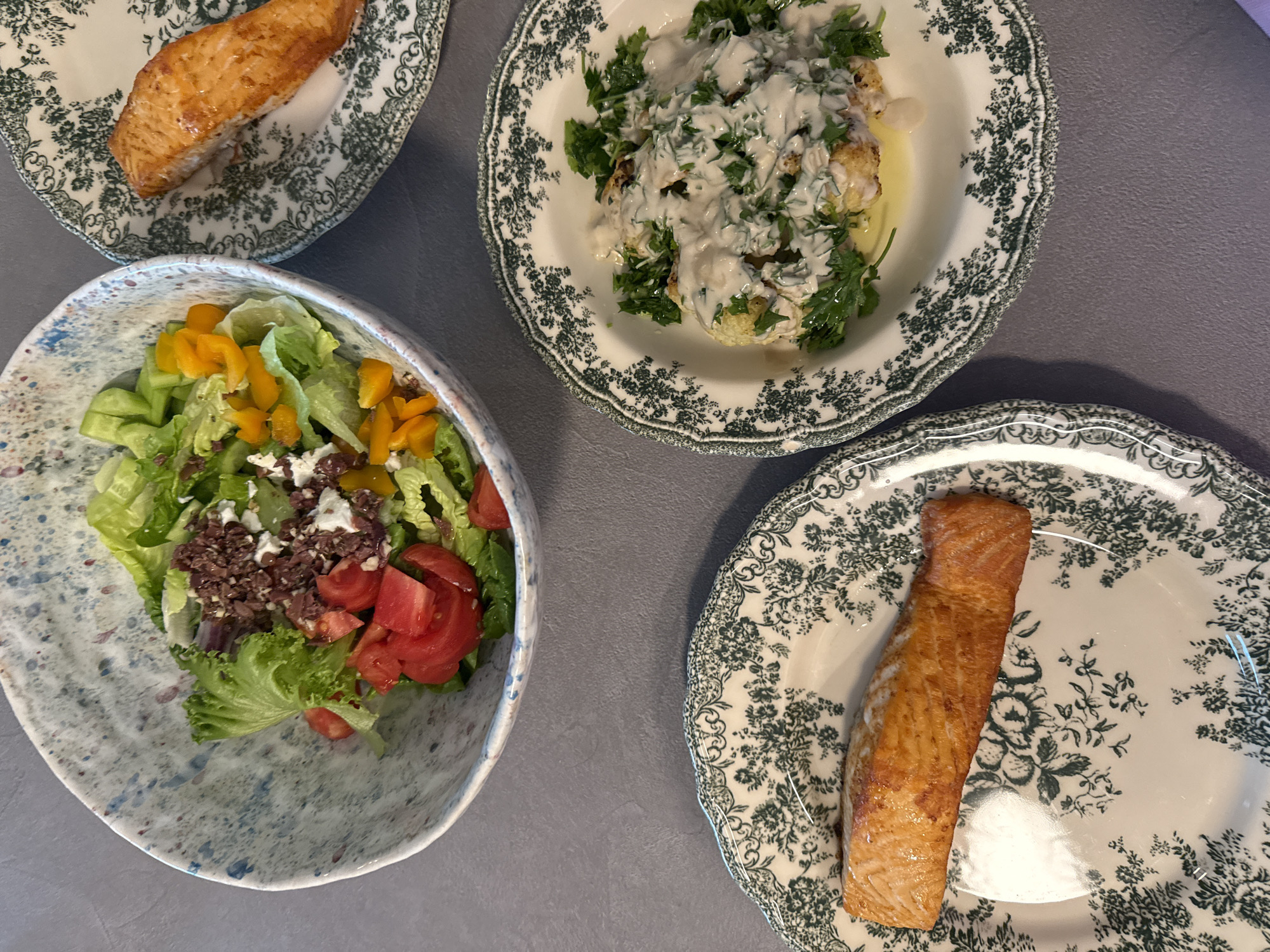 The image shows four dishes of food arranged on a gray surface. Two plates contain salmon, one plate contains cauliflower covered in a white sauce and parsley, and the fourth dish is a bowl of salad with lettuce, tomatoes, olives, peppers, and feta cheese. The plates have a green and white floral pattern. There are no people visible. The location is unknown and could not be determined from the image.