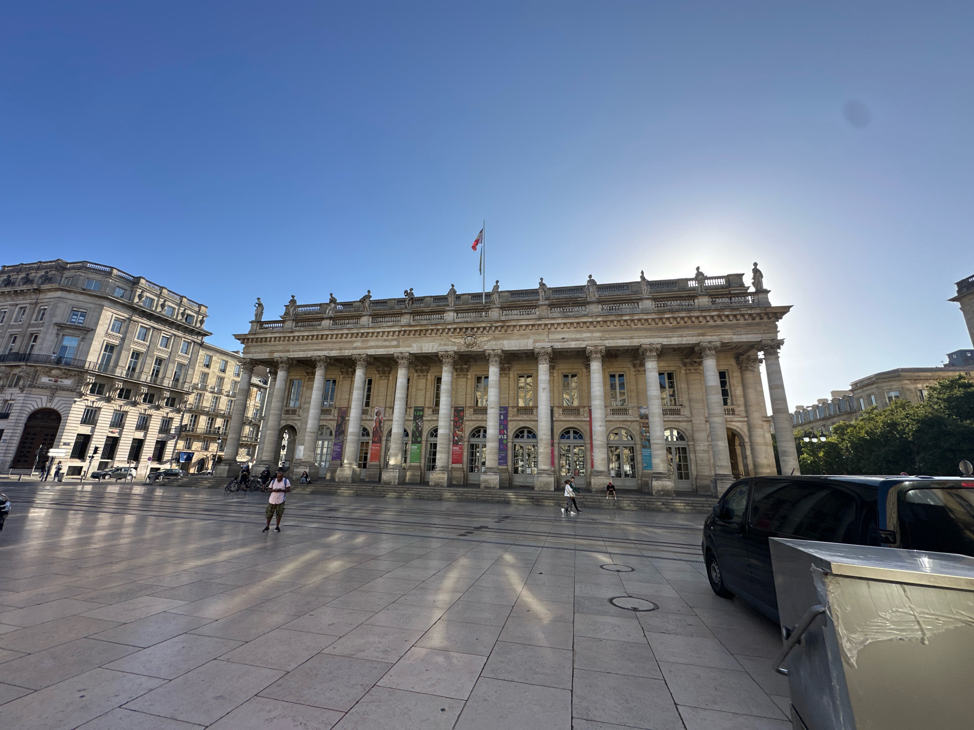 The image captures a wide view of the Place de la Comédie in Bordeaux, France. The Grand Théâtre de Bordeaux, a magnificent neoclassical building with a grand colonnade and a row of statues along its roofline, dominates the center of the frame. The French flag flies from a flagpole atop the building. Sunlight streams from the right, casting long shadows across the large, paved square. Several people are visible on the square, including a woman standing in the middle ground wearing a light-colored shirt and patterned shorts, and a couple walking in the foreground. A black van is parked on the right side of the square. Other buildings, including a lighter-colored building to the left of the Grand Théâtre, line the perimeter of the square. The sky is clear and blue.