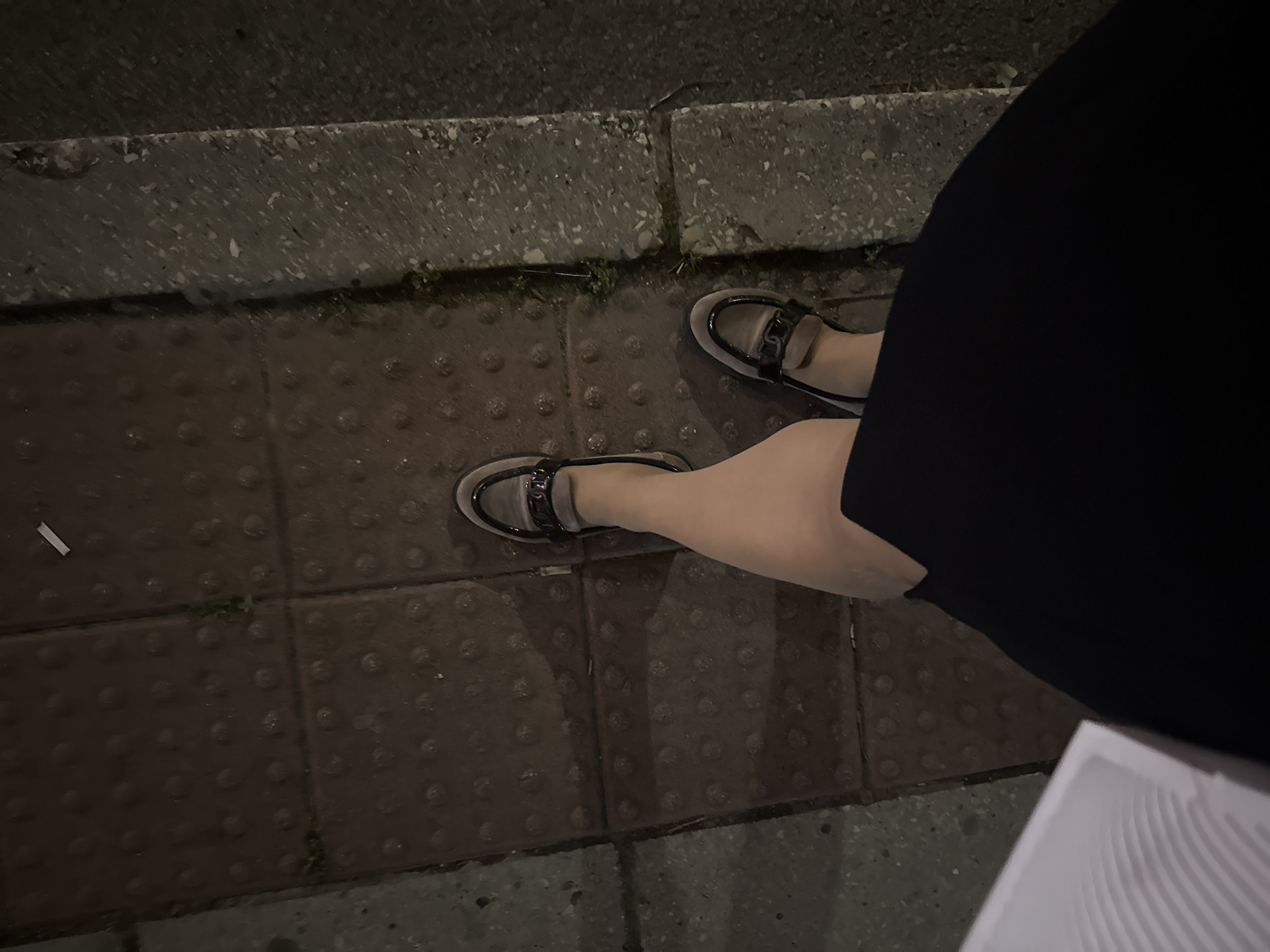 The image presents a high-angle, overhead perspective of an individual's lower body standing on a sidewalk in dimly lit conditions in Unknown, Unknown. The person is wearing a dark, likely black, skirt or dress, skin-toned hosiery, and light brown or beige loafers with black trim and a decorative metallic chain-link element across the vamp. The sidewalk is composed of dark reddish-brown square tiles, some exhibiting raised circular tactile bumps. Minor vegetation and dirt are visible within the tile seams. To the right, a textured concrete curb demarcates the sidewalk from an adjacent darker surface. A white rectangular object, consistent with a cigarette butt, is located on the sidewalk in the upper right portion of the frame. A partially visible white object with a ribbed surface, resembling a disposable container, is present in the lower left corner. Shadows are cast by the legs and shoes, indicating an off-center light source.