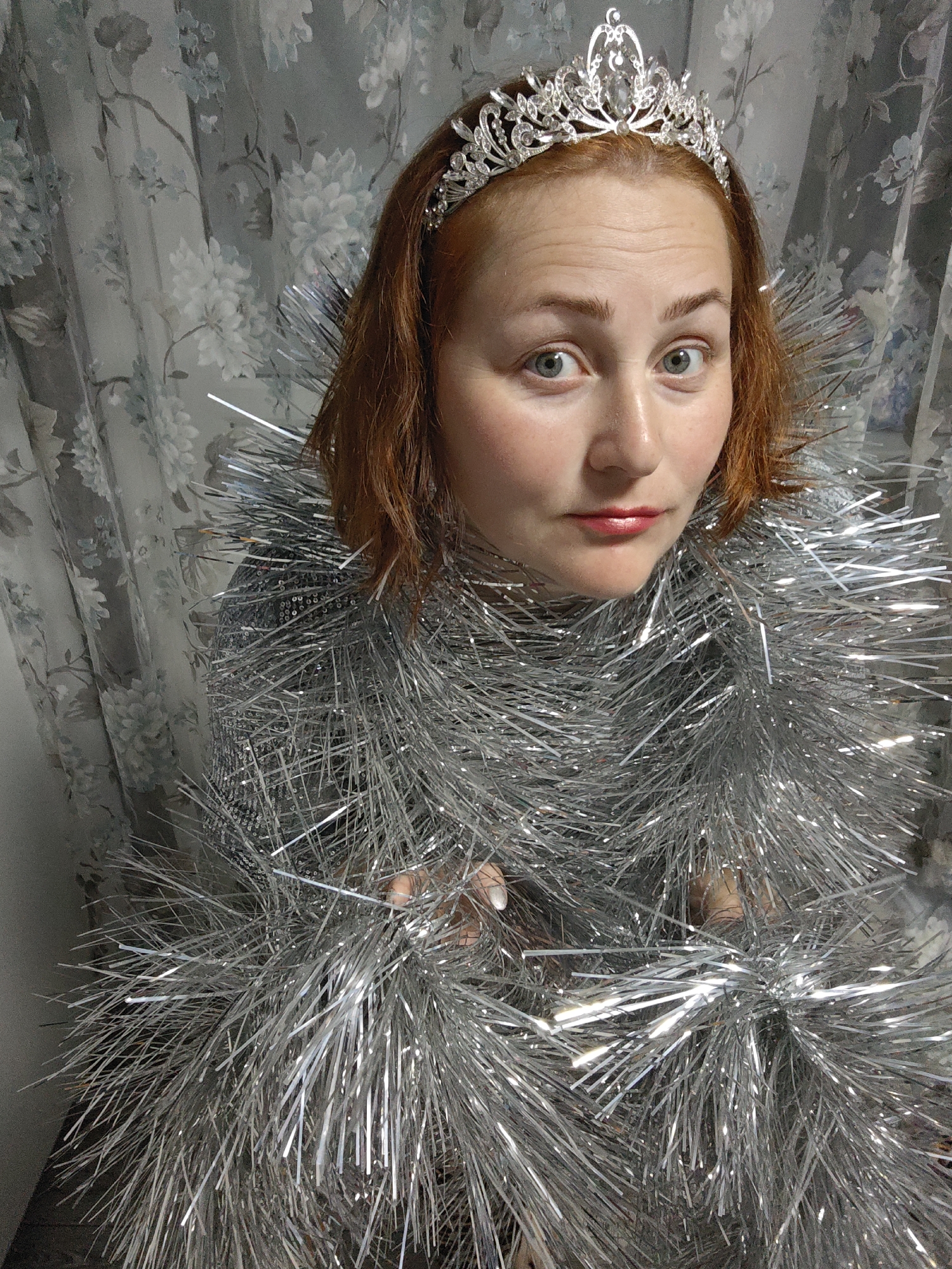A woman with red hair and fair skin is the central subject of this image. She is wearing a silver tiara on her head and is draped in silver tinsel, which obscures much of her clothing and torso. Her expression is one of slight surprise or concern, with wide eyes and a subtle upward tilt of her head. Red lipstick is visible. The background consists of a patterned curtain with floral motifs in shades of gray and white. The lighting appears to be somewhat diffused, creating soft shadows. Based on the description of the location as Markovskii, Russia, this scene is likely taking place indoors, in a home or apartment.