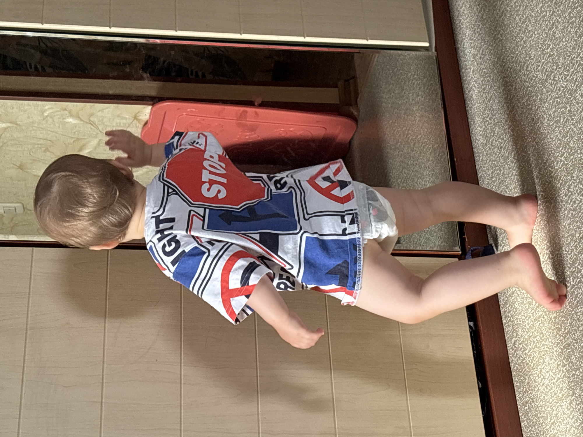 The image shows a young child, viewed from the back, standing in a room with wooden doors and a mirrored surface. The child is wearing a t-shirt with a graphic design featuring road signs, including a prominent red "STOP" sign, and a diaper. The child's right arm is raised. The environment appears to be indoors, with a carpeted floor. There is no visible indication of the specific location within Naberezhnye Chelny, Russia.