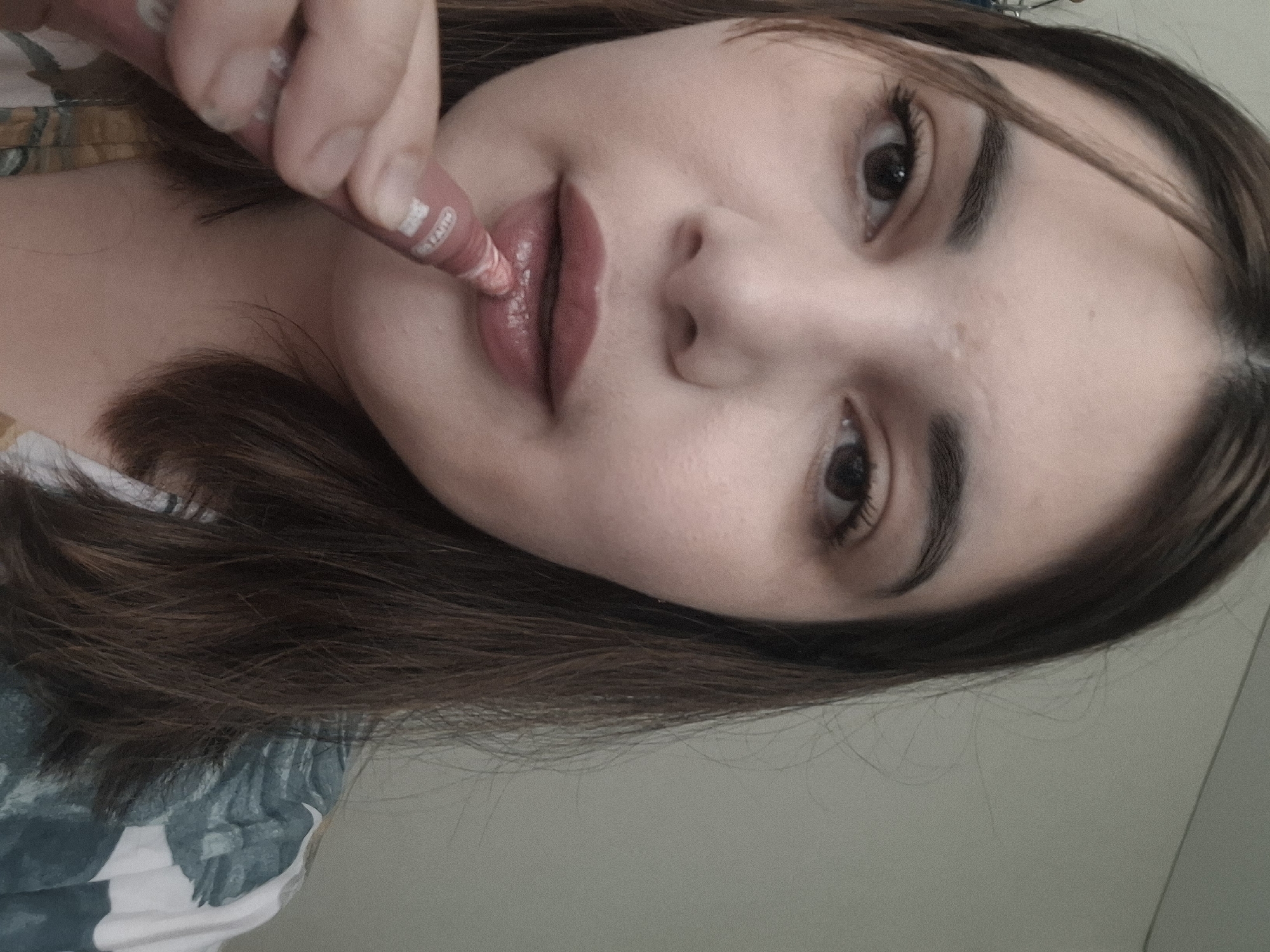 A close-up, slightly tilted, selfie shows a woman applying lip gloss. The woman has light skin, brown hair, and brown eyes. She is wearing a floral-patterned top. In her right hand, she holds a pink tube of lip gloss. The location is unknown and there is no indication it is in Амзя, Russia.