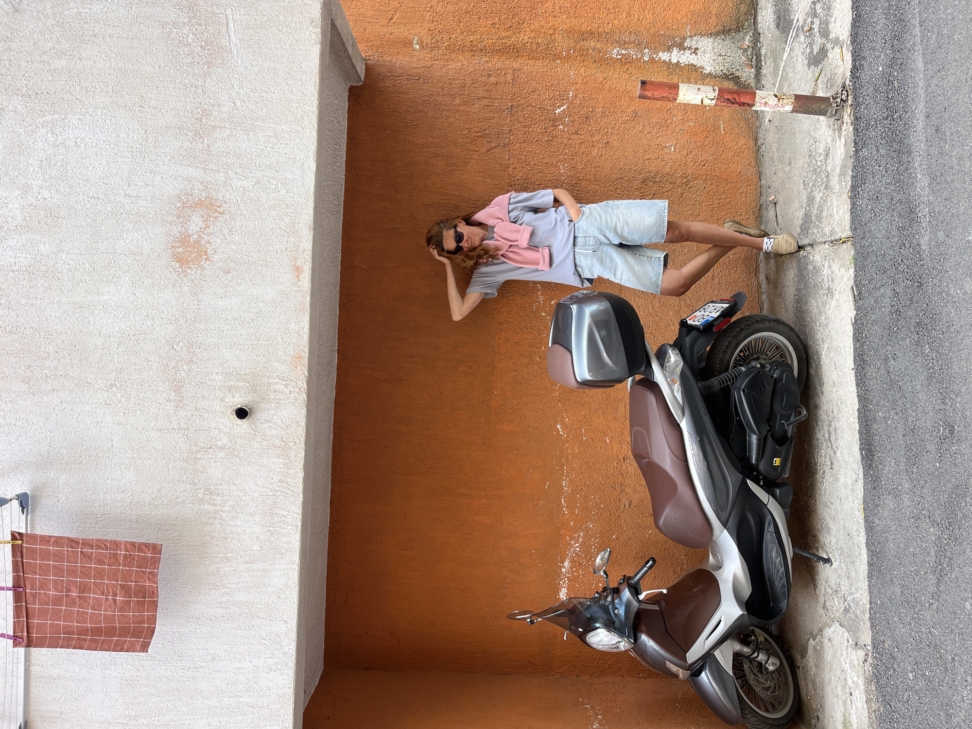 A full-length, eye-level image rotated 90 degrees clockwise shows a man lying against an orange wall next to a parked moped. The man wears sunglasses, a grey shirt, light-wash denim shorts, and tan shoes; a pink sweater is draped around his neck. He is posed propped on one arm. To the left of the wall is a white wall with a dark hole and a towel on a rack. The moped is parked on the sidewalk. The moped has a silver top case and a brown seat. A red and white striped pole is visible near the man's feet. The location is identified as Budva, Montenegro.