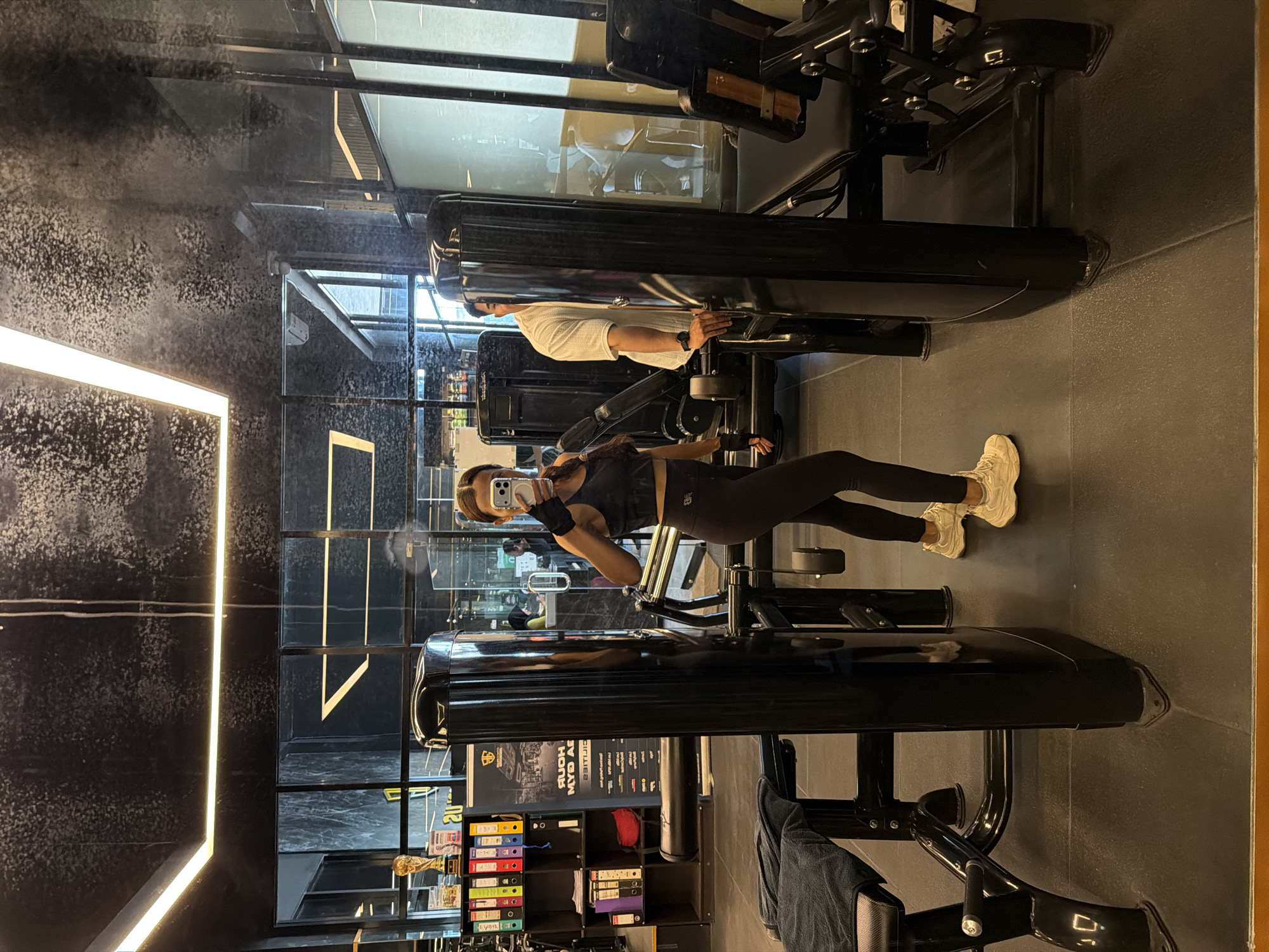 A woman in a black athletic outfit stands in front of a mirrored wall in a gym in Denpasar, Indonesia, taking a selfie with her smartphone. To her right, a man wearing a white shirt is using a weight machine. The gym contains various exercise machines and storage shelves filled with binders. The floor is tiled in a dark color.