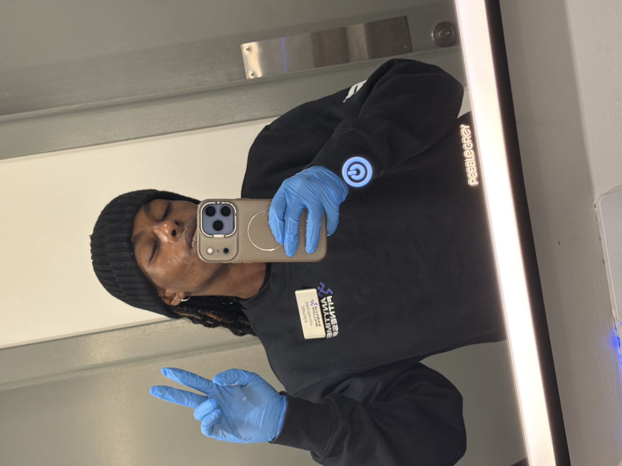 A person is seen in a mirror selfie, captured from a slightly elevated and tilted perspective. The individual, appearing to be male, is wearing a black beanie and a black long-sleeved shirt. They are also wearing light blue disposable gloves on both hands. Their eyes are closed, and their mouth is slightly open, suggesting a relaxed or playful expression. They are holding a smartphone with a light-colored case (featuring a triple-lens camera array) to take the photo, which is visible as a reflection.
With their left hand (gloved), the person makes a "peace sign." On their right sleeve, a prominent glowing blue circular logo resembling a power symbol is visible. A white name tag or badge is pinned to their shirt, reflected in the mirror.
The setting appears to be indoors, possibly a bathroom or changing room, indicated by a prominent vertical strip of bright light on the right side of the mirror and a metallic fixture above.
Visible text includes:
* On the mirror, reversed: "PEBBLEGREY"
* On the name tag/badge, reversed: The purple "ANYTIME FITNESS" logo (a running man) and the name "Kamaal" (partially legible).
* On the right sleeve: A glowing blue power-on/off symbol.