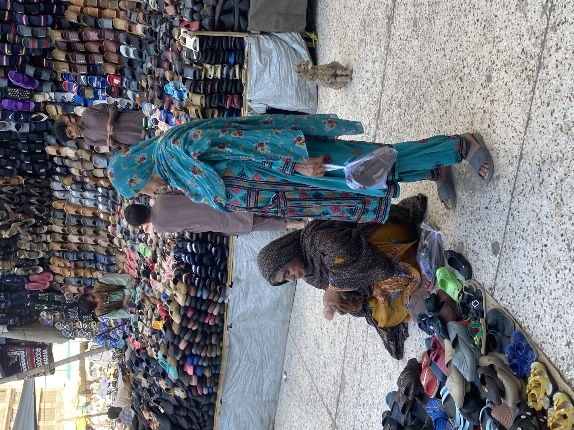 A medium-angle outdoor shot captures a crowded marketplace scene in Quetta City Tehsil, Pakistan. The view is slightly tilted.
A woman in a turquoise floral dress and head covering stands, holding a dark-colored bag. Behind her, a man in a brown shirt stands near rows of sandals displayed for sale. To the right, another woman in a dark floral head covering and a yellow garment bends over a collection of sandals. A cat sits on the ground near a draped surface.
Rows of sandals line the backdrop. Other shoppers and vendors are visible in the background. The ground is a textured gray. Buildings are visible in the far background under a slightly overcast sky.