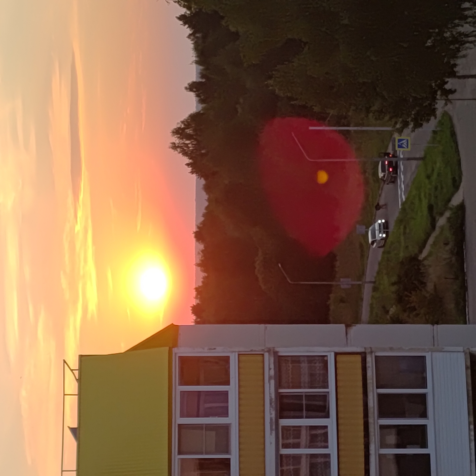 The image captures a sunset view from an apartment building in Марковский, Russia. In the foreground, the side of a multi-story building is visible, featuring yellow and white facades with multiple windows and balconies. To the right, a dense line of trees forms the backdrop, with a hazy horizon visible beyond them. A road curves through the scene, with two cars present: a white car in motion on the left side of the road, and a darker car stopped near a pedestrian crossing on the right. The sky is filled with warm orange and yellow hues, characteristic of a sunset, with scattered clouds. A prominent red lens flare is present in the mid-ground, obscuring some details of the road and trees. The overall impression is a calm, suburban evening scene.