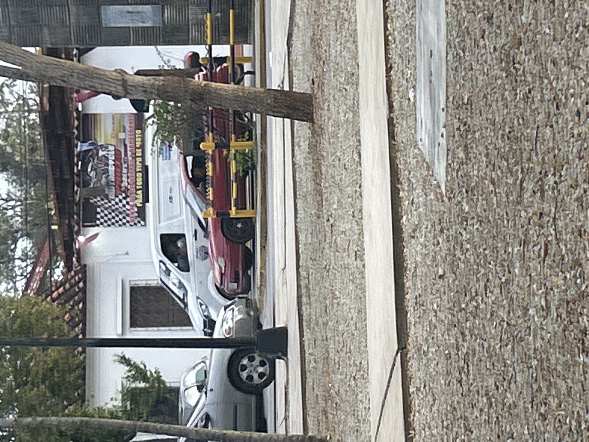 The image captures a street scene with a variety of vehicles and buildings, possibly in Santa Ana, Venezuela. A light-colored sedan is parked on the left side of the frame, partially obscured by a tree. To its right, a white van is stopped, with a driver visible inside. In the foreground, a red car is positioned in the middle of the road, with its front facing towards the right. The car has a person behind the wheel and what appears to be a red motorcycle passenger behind it. Behind the vehicles, a building with a white facade is visible, featuring a checkered flag design and a banner with text that is difficult to read. The area is paved with a textured surface, and there are yellow and black striped bollards lining the road. The environment suggests a typical urban or semi-urban setting with trees and structures indicating human activity.