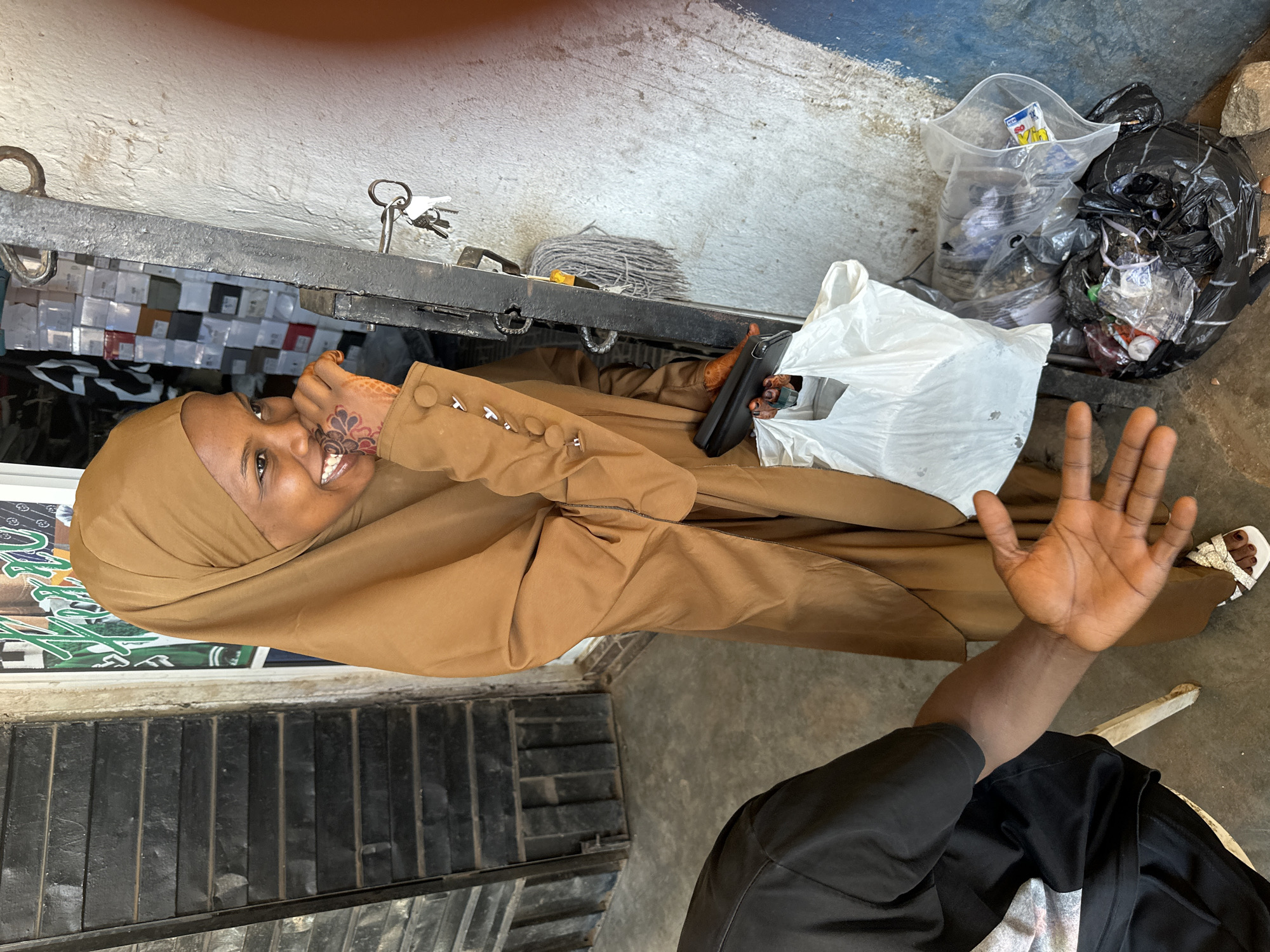 In Bukuru, Nigeria, a young woman wearing a brown hijab and matching outfit smiles widely as she holds a white plastic bag and a dark-colored object, possibly a wallet or small pouch. Her left hand, adorned with henna, is brought to her mouth, and her right hand is visible, extended outward. She stands near a metal gate with keys attached. To her left, a person in a black t-shirt is partially visible, their arm and open hand also in view. The ground is concrete, and to the right, several trash bags are piled up. Behind the woman, shelves stocked with various items are visible through an open doorway, suggesting a shop environment.