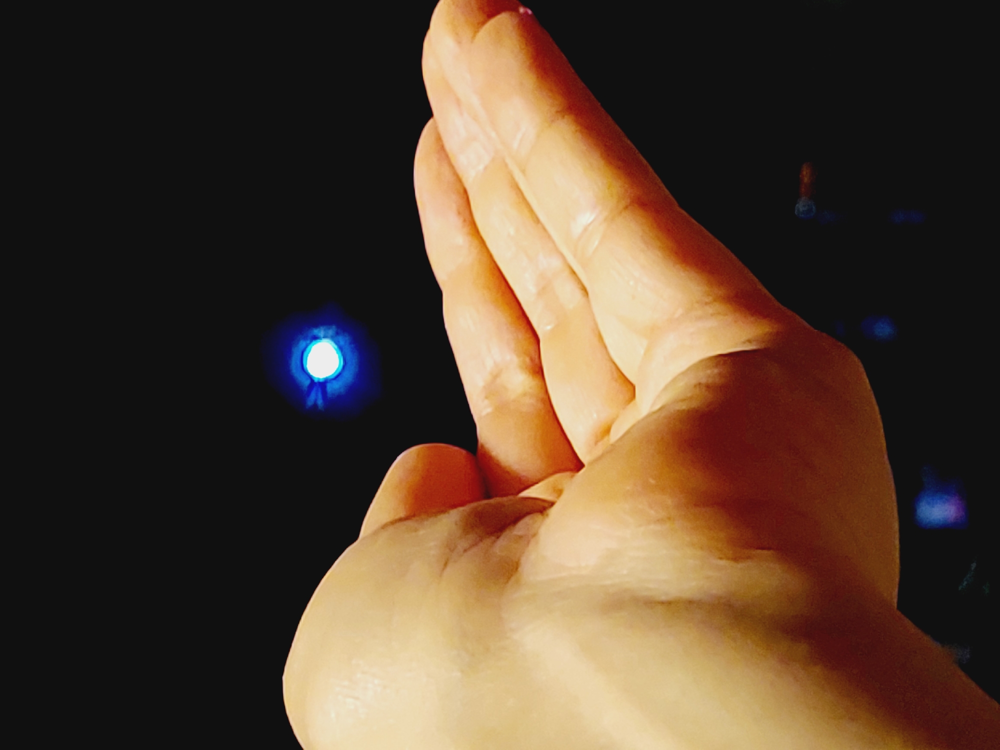 A close-up shot shows a person's hand in the foreground, palm open and facing upward, with fingers slightly curled. In the blurred background, a small, bright blue light source is visible against a black backdrop. The scene appears to be captured in low light conditions, suggesting an indoor or nighttime setting, and is not specific to Sozopol, Bulgaria.
