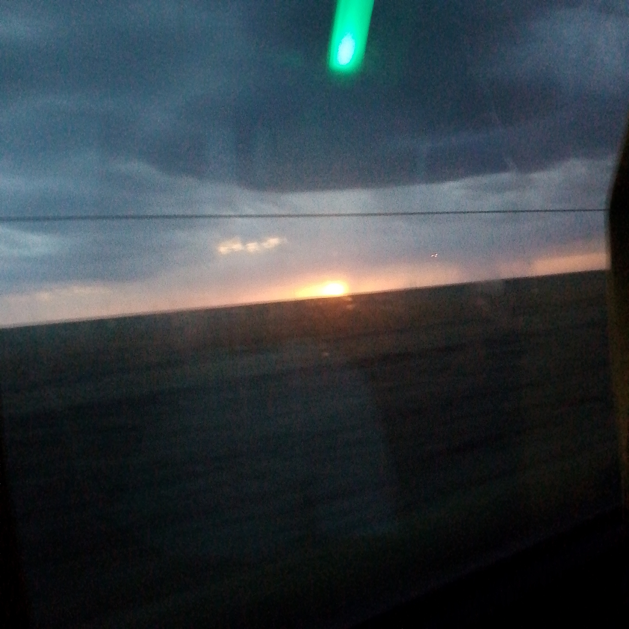 The image captures a twilight scene, likely viewed from a window, given the presence of a faint green light source at the top and what appears to be a reflection or smudge on the glass. The horizon is marked by a faint line, separating a dark, blurry landscape from the sky. The sky itself transitions from a deep blue-gray at the top to lighter, softer tones near the horizon, with subtle hints of clouds. A bright, diffused light, suggestive of the setting sun, is visible low on the horizon, casting a warm glow on the lower part of the sky. The landscape below is predominantly dark and indistinct, suggesting it is either nighttime or heavily shadowed. There are no discernible people or specific structures visible in the scene. Without additional context or visual cues, it is impossible to definitively place this image within the city of Adyk, Russia.