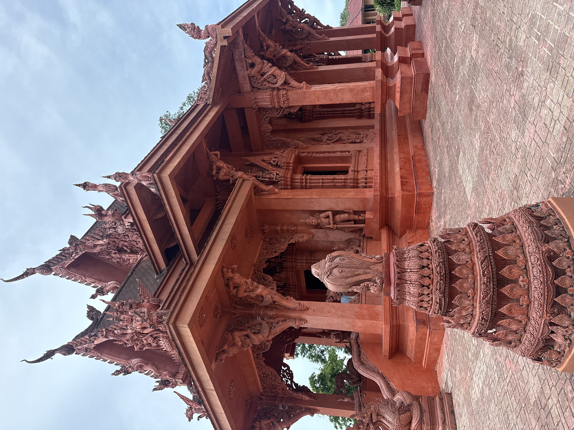 This is a full, eye-level, outdoor shot of the exterior of the Sanctuary of Truth in Ko Samui District, Thailand. The building is constructed of wood and is painted a bright red. The building has a multi-tiered roof with intricate carvings and a central spire. The building is surrounded by a paved courtyard. In the foreground, there is a decorative pillar with a tiered base and intricate carvings. The sky is cloudy and overcast. There are no people visible in the image.