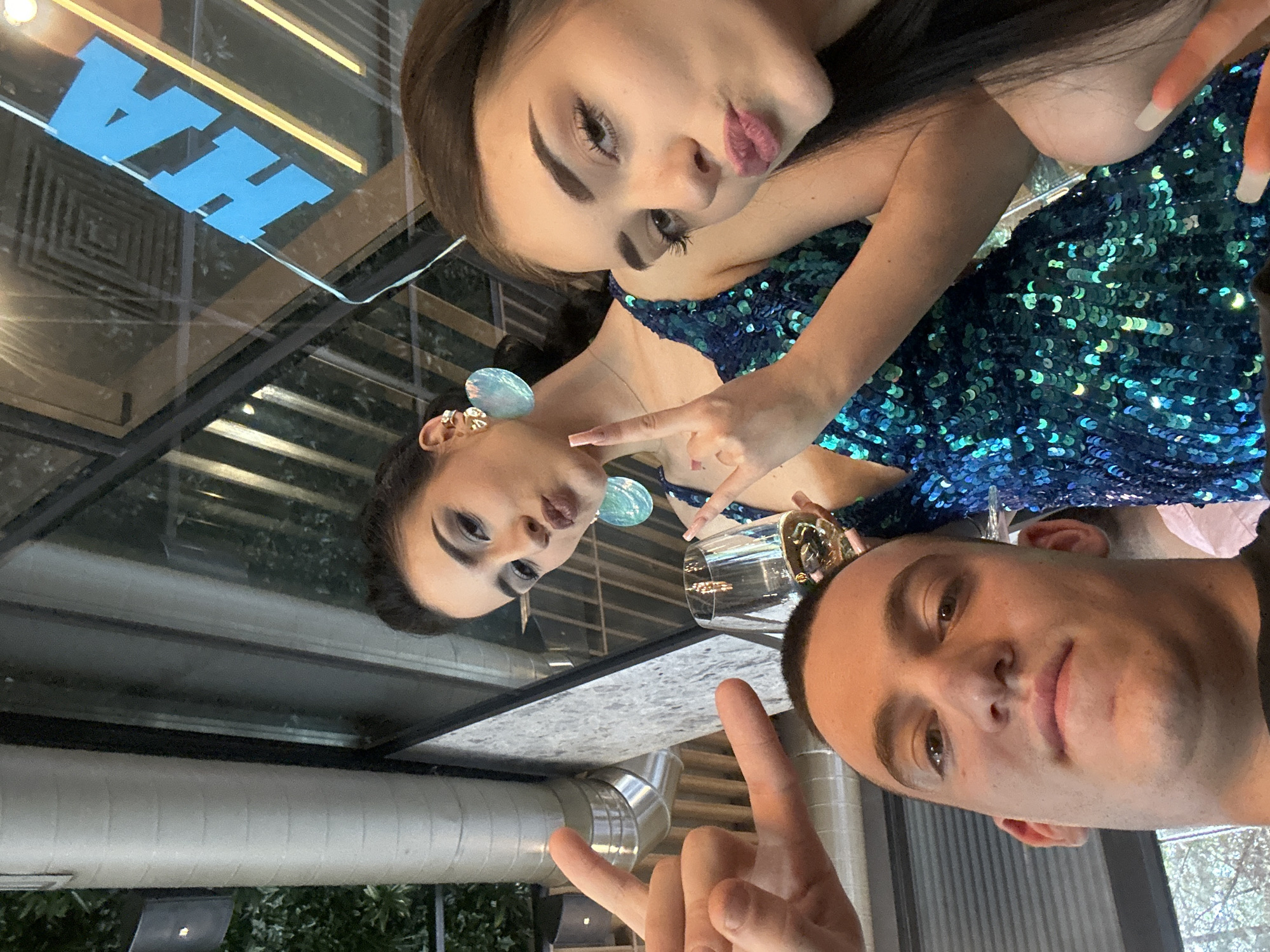 A selfie taken indoors, possibly in a restaurant, features two women and a man. One woman, with long brown hair and wearing a shiny blue dress, is making a kissing face. The second woman, reflected in a surface, has dark hair pulled back and wears large, round earrings. The man is positioned in the foreground, partially visible. The background includes a partial view of what appears to be a window or reflective surface with blue lettering. There is also a drinking glass and a metallic object in the vicinity. Outside, greenery is visible through the window. It cannot be determined where this image was taken with the information provided.